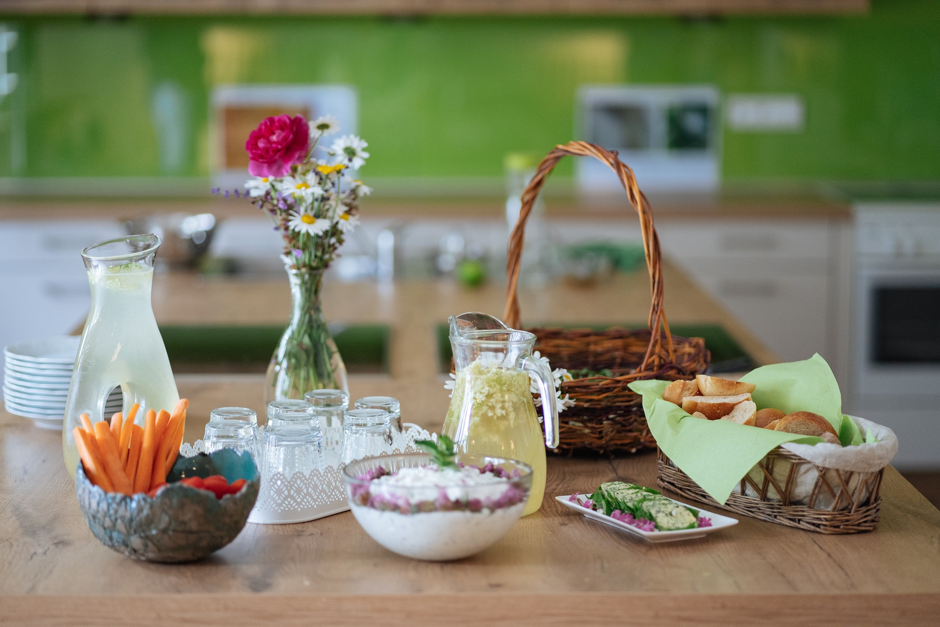 Une table avec un bouquet de fleurs colorées, un panier rempli de pain, une limonade à base de sureau, un fromage blanc naturel affiné aux herbes, plusieurs verres et un bol de légumes frais.