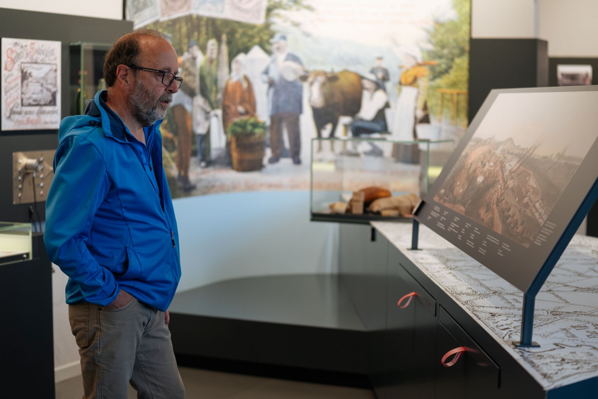 Un homme regarde une exposition dans un musée. Il est debout, vêtu d'une veste bleue et d'un pantalon gris, les mains dans les poches.