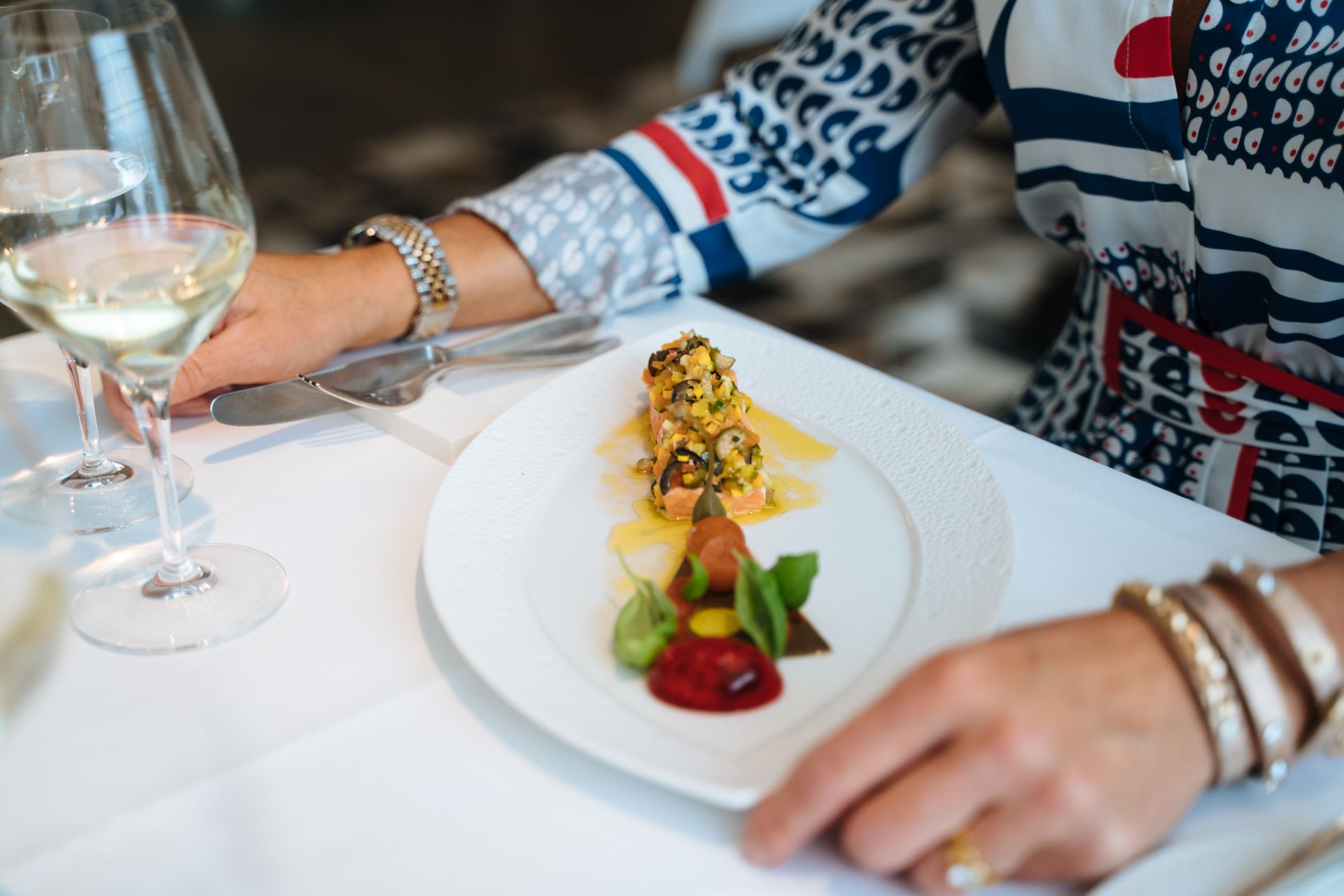 Le point central de cette photo est une assiette de saumon avec des garnitures et une petite salade d'accompagnement sur une assiette blanche, avec un verre à vin et les mains d'une invitée visibles au bord de la photo.