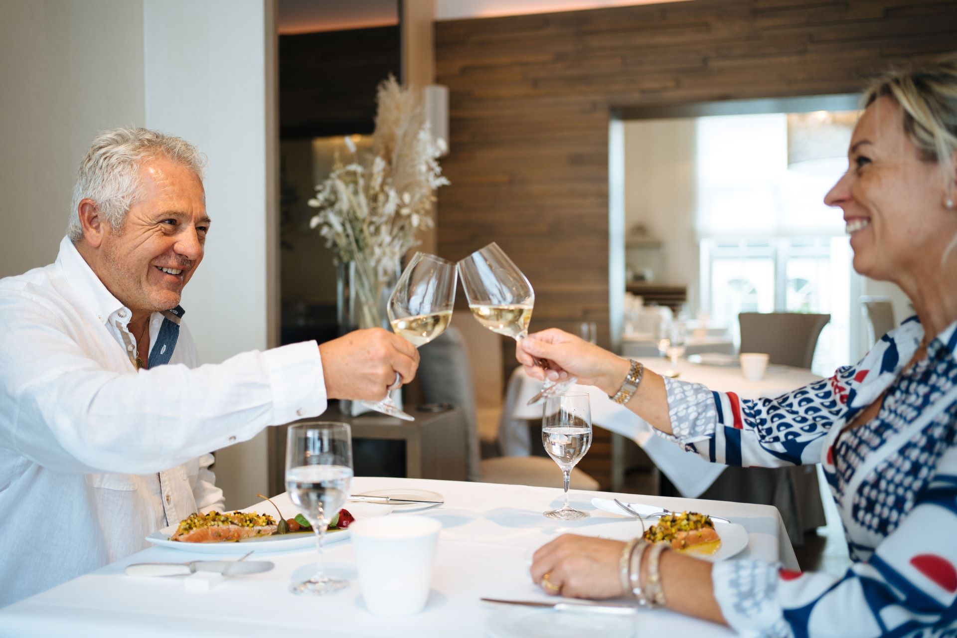 Un homme et une femme assis à une table chic avec une nappe blanche et des assiettes blanches trinquent avec leurs verres à vin alors qu'ils s'apprêtent à manger du saumon.