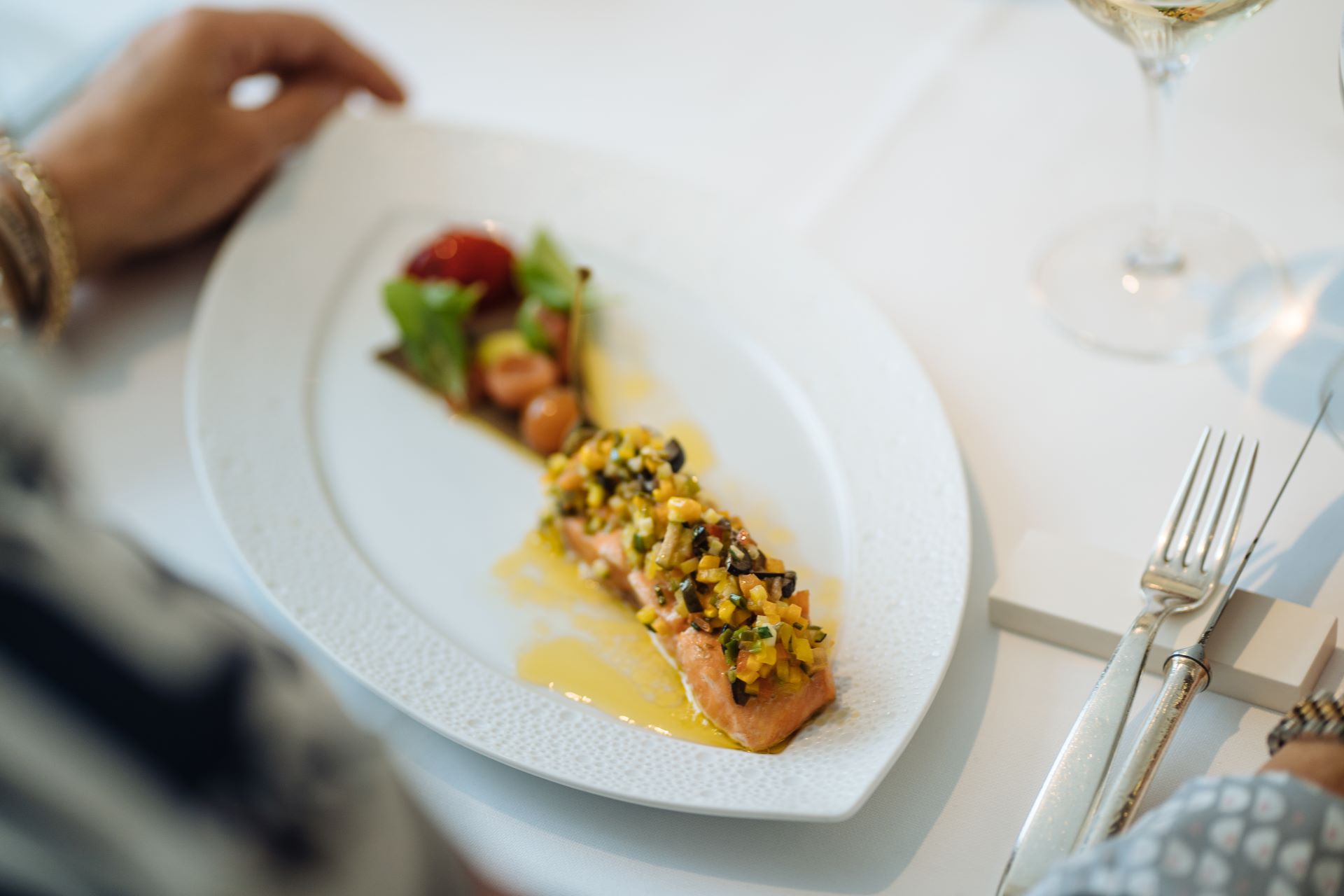 Une assiette de saumon avec des garnitures et une petite salade d'accompagnement est au centre de cette image. La main d'un invité est visible, mais hors champ, à gauche de l'assiette, qui est posée sur une nappe blanche propre.