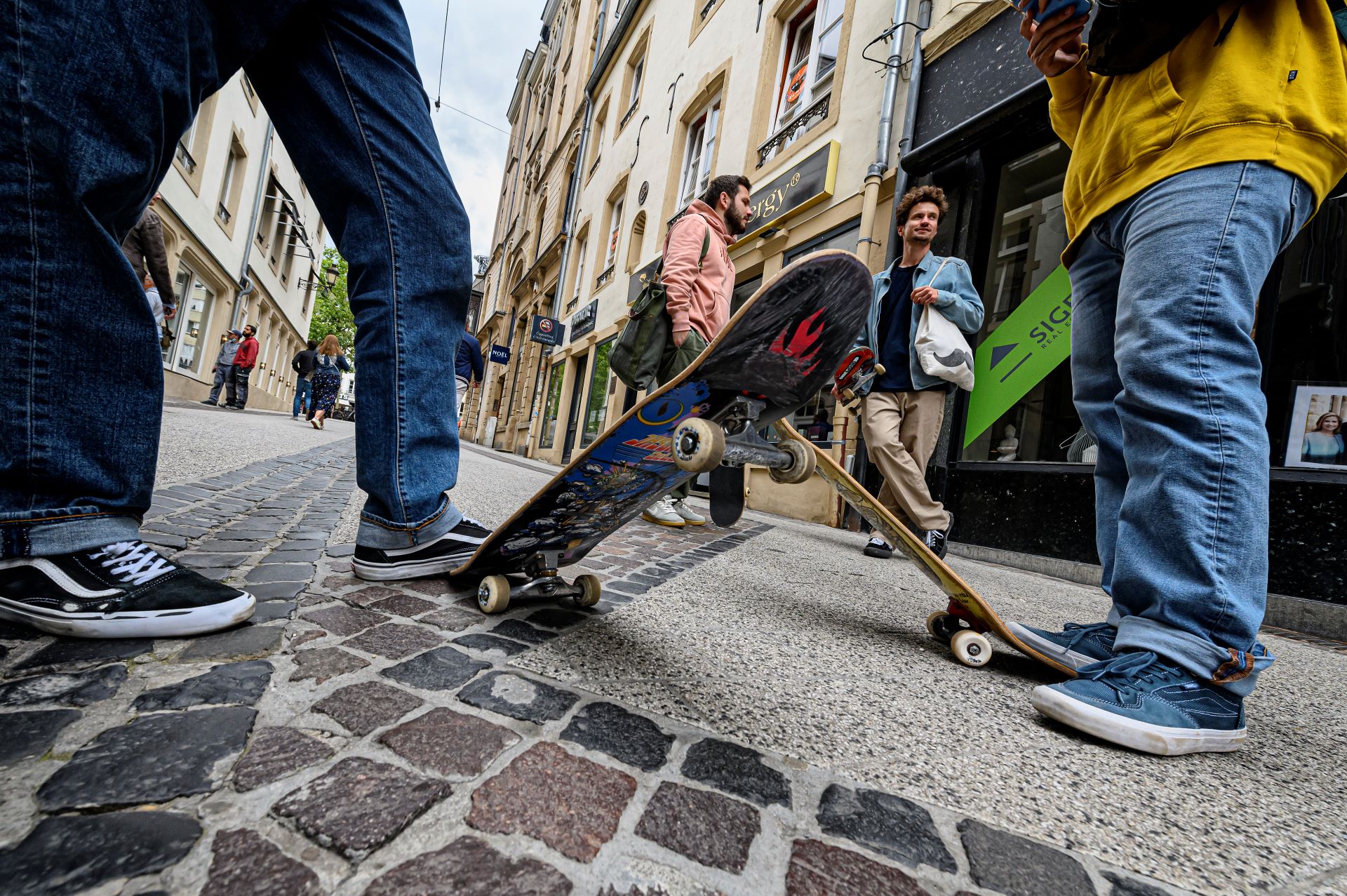 Des skateurs discutent dans une rue du centre ville de Luxembourg. Deux d'entre eux sont hors champ, mais on peut voir leurs skateboards au premier plan.