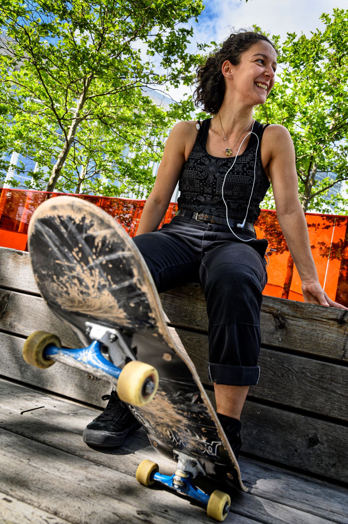 Une femme est assise, souriante, sur un banc et regarde vers la gauche. Son pied est posé sur un skateboard, qui se trouve au premier plan de la photo.