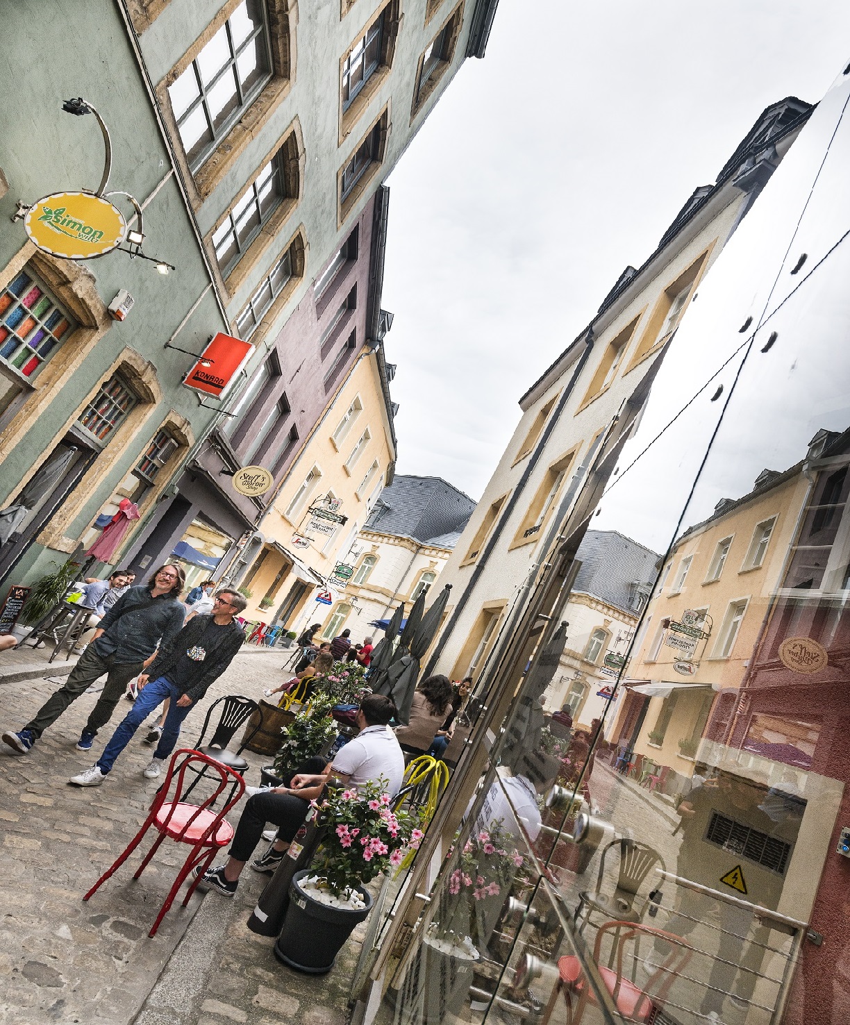 Une ruelle dans la vieille ville de Luxembourg avec des maisons et des cafés colorés. Deux hommes traversent la ruelle. Un homme est assis devant un café.