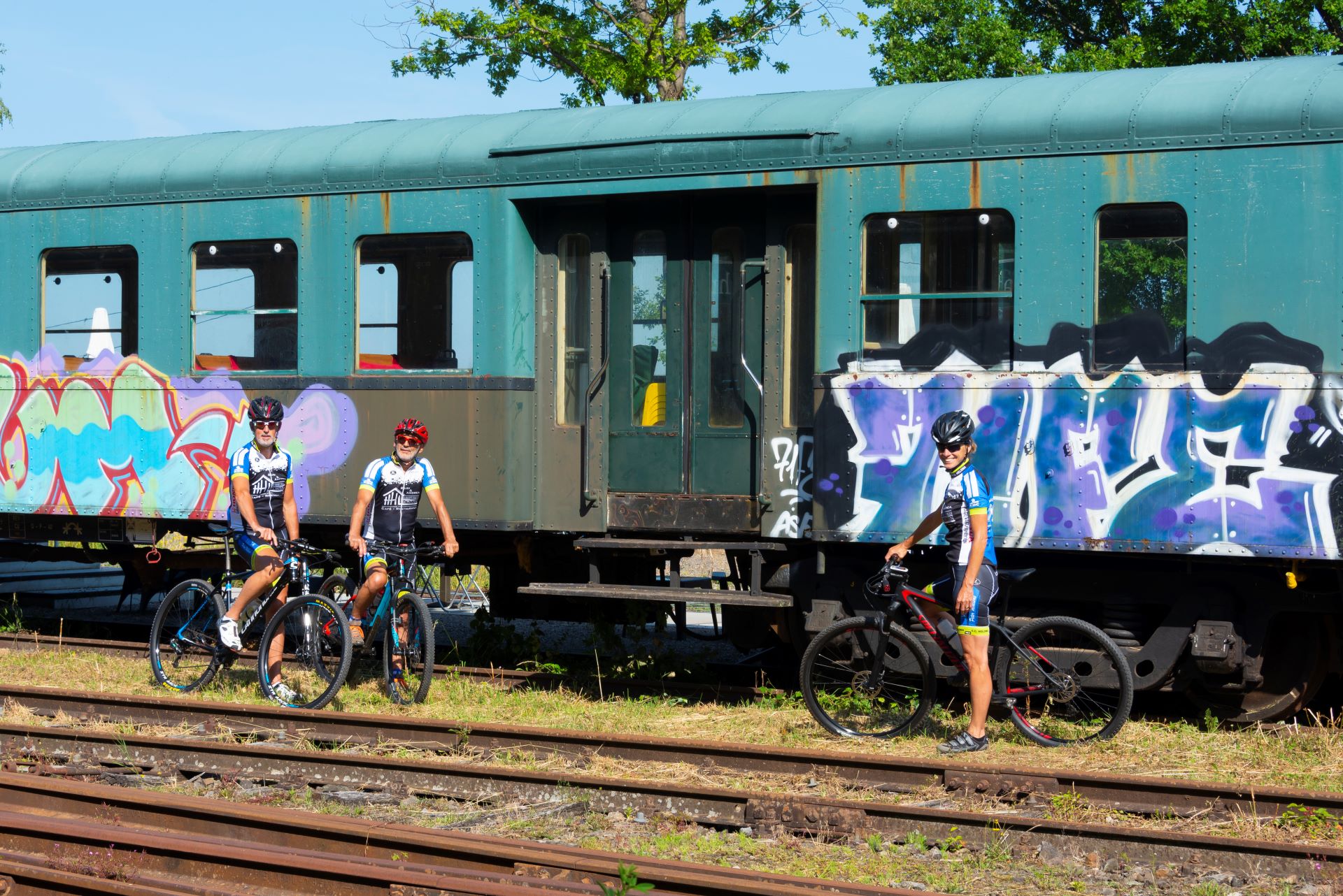 Trois vététistes se tiennent devant un train peint à la bombe de couleurs vives. Les rails sont visibles au premier plan.