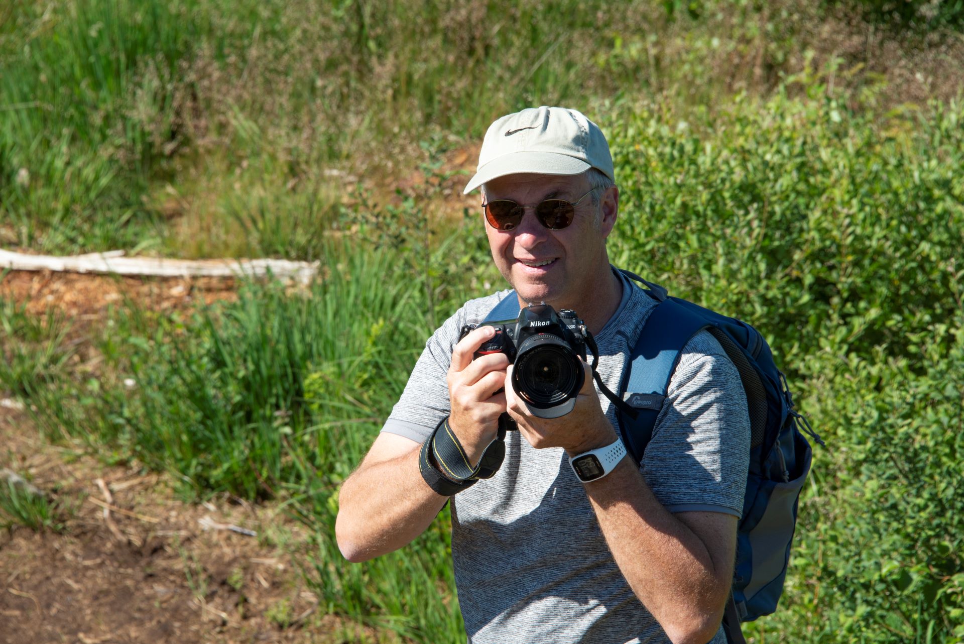 Le guide touristique est photographié regardant l'appareil photo et souriant. Il porte sa caméra dans ses mains et la dirige vers le spectateur.