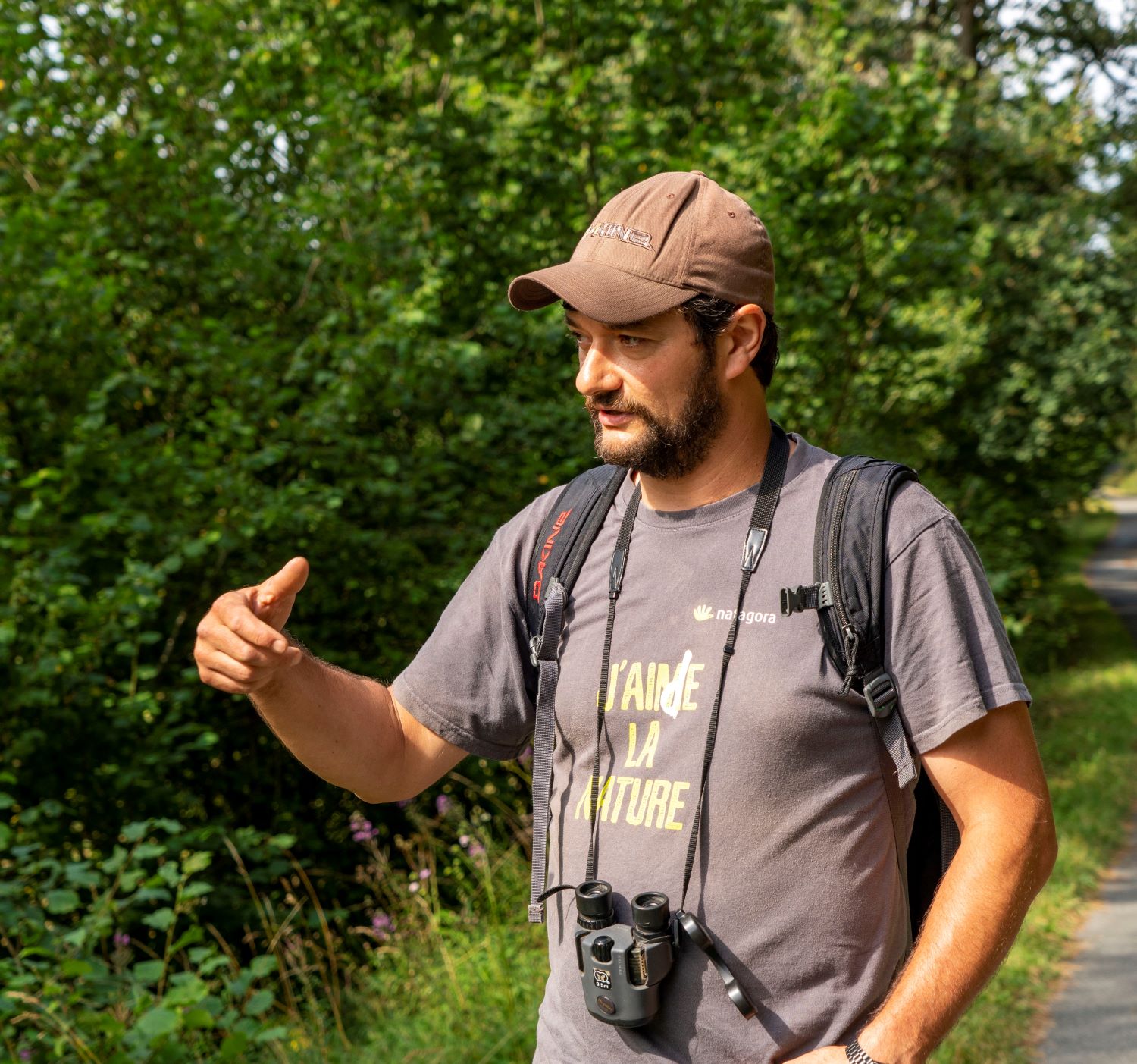 Un homme portant une casquette marron et une chemise avec les mots "J'aime la nature" fait des gestes en parlant de la nature sauvage de la région. Un appareil photo est suspendu à son cou.
