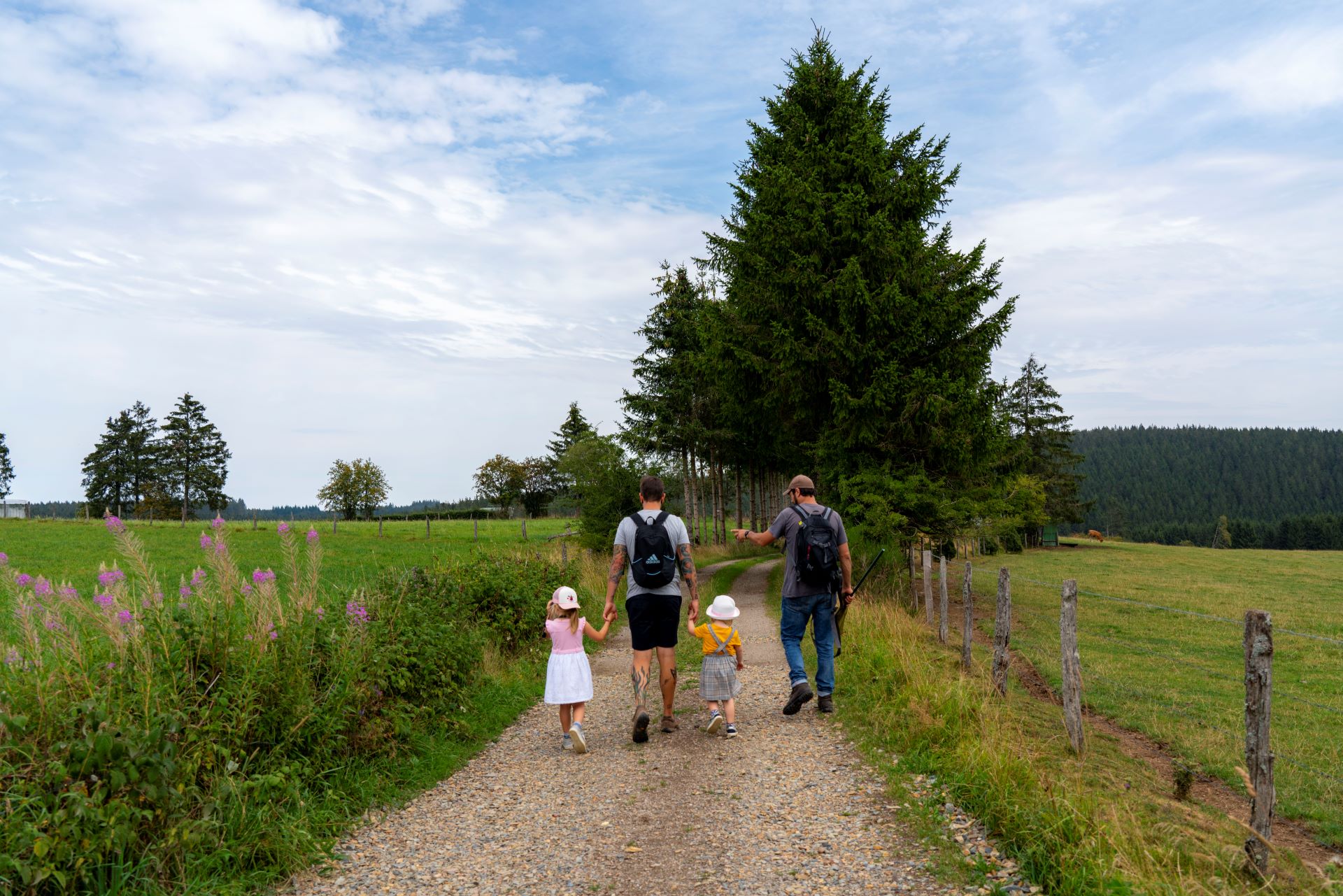Un homme tient les mains de ses deux petites filles alors qu'elles marchent à côté d'un guide de la nature. Le chemin est large et caillouteux, la nature environnante et les arbres sont verts.