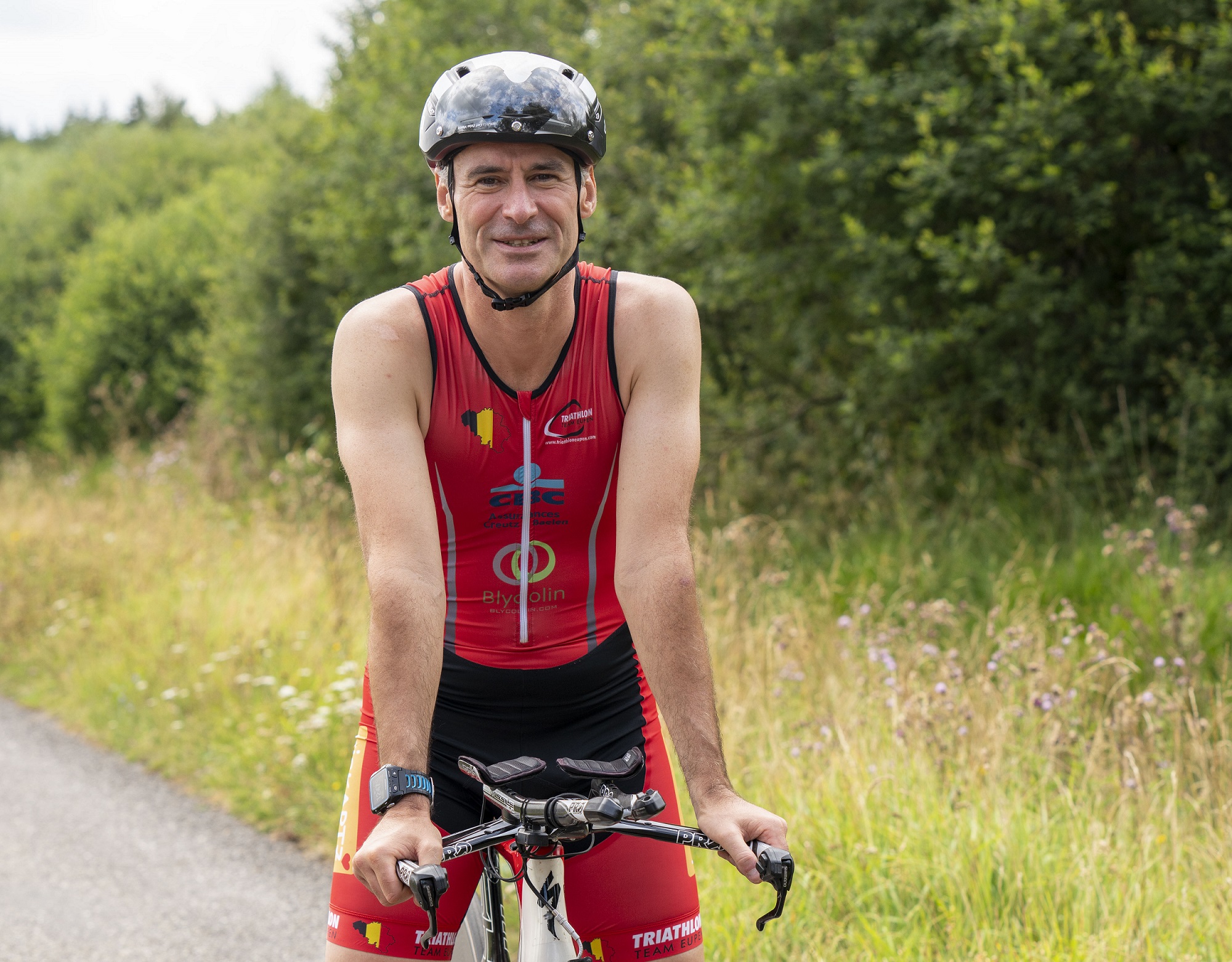 Photo portrait d'Oliver Henz sur son vélo de course. Il regarde de face la caméra et sourit. Il porte sa combinaison de triathlon noire et rouge et un casque noir.