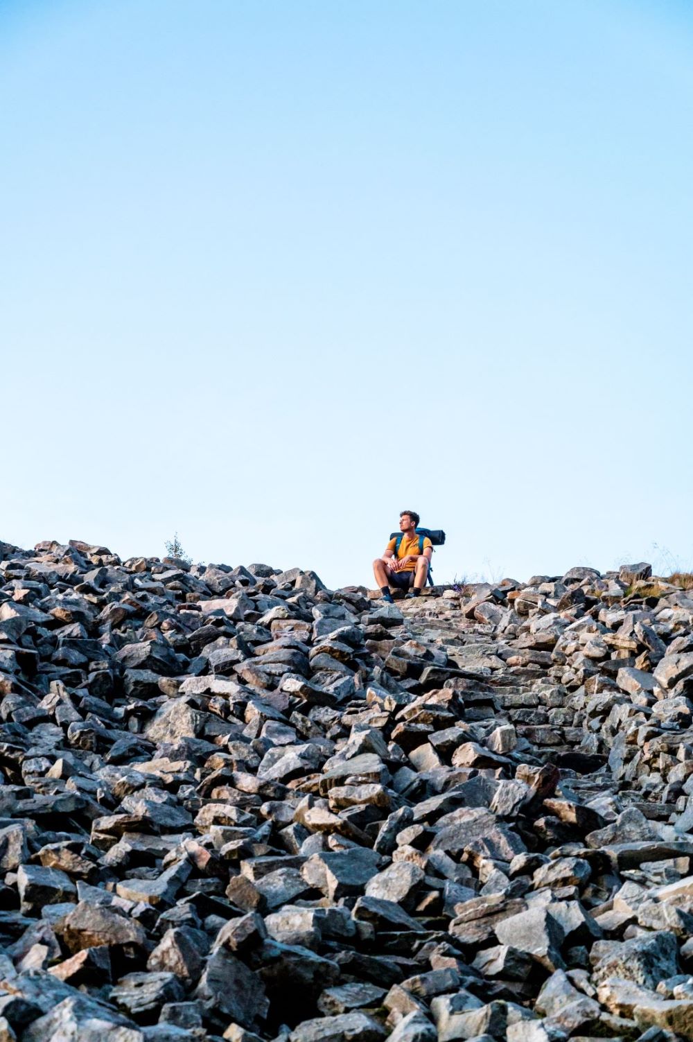 Un homme portant une chemise jaune et un équipement de randonnée est photographié au sommet d'un tas de rochers.