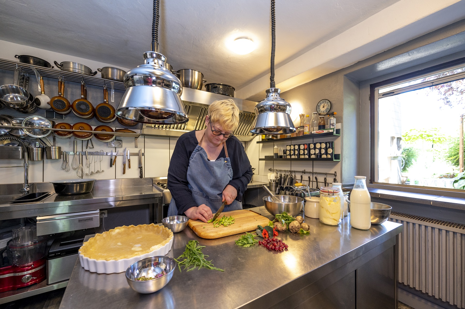 La cuisine de la maison de campagne des Spanier. Anja Faust-Spanier se tient debout devant l'îlot de cuisson en métal et coupe des légumes. Le moule pour la quiche est là et les ingrédients sont étalés sur le buffet. En arrière-plan, vous pouvez voir la cuisine avec des pots et des épices.