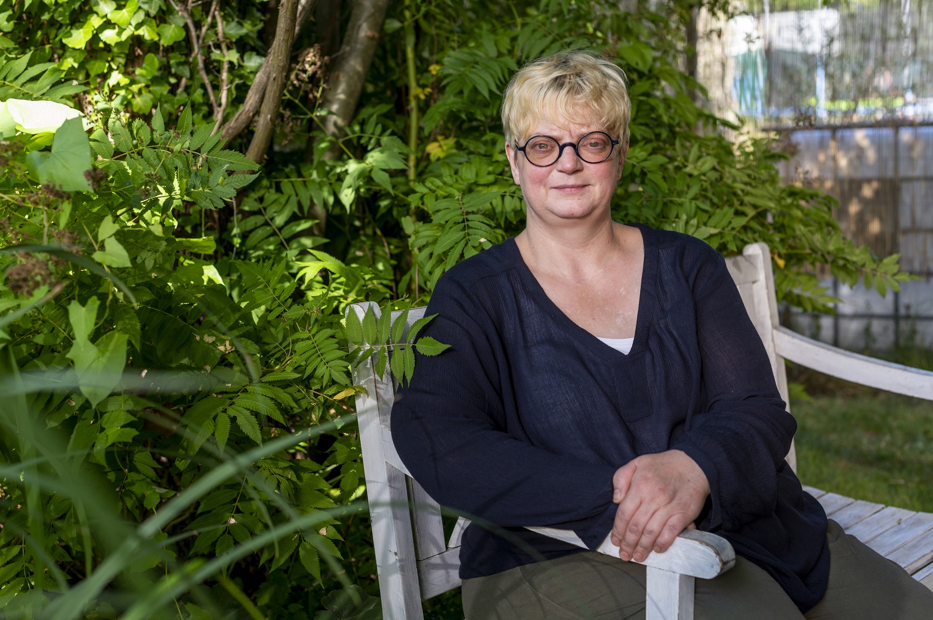Anja Faust-Spanier, chef et propriétaire de Landhaus Spanier, est assise sur un banc blanc. Elle porte des lunettes rondes noires, un chemisier noir et un pantalon vert foncé. Il y a des plantes vertes en arrière-plan.