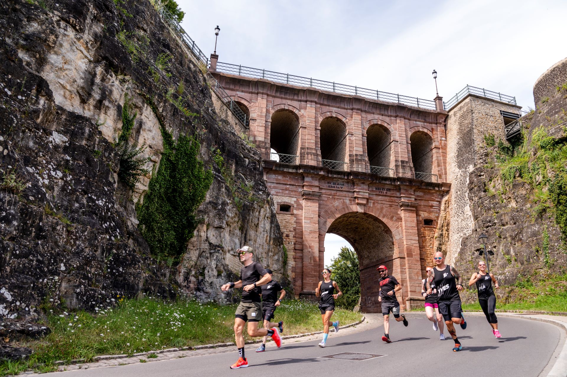 Sept membres du groupe de coureurs FatBetty.run émergent de sous un vieux pont de pierre au Luxembourg. Ils sont habillés de vêtements de course noirs et courent sur une route pavée.