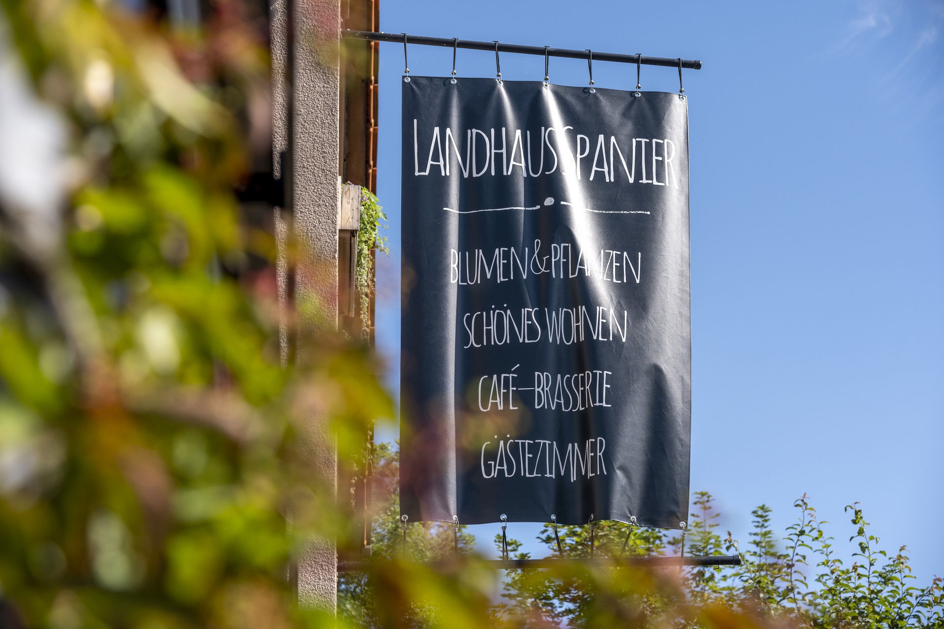 Le drapeau sur la maison de Landhaus Spanier avec les mots suivants : Fleurs et plantes, Belle vie, Café-Brasserie, Chambre d'hôtes. Au premier plan, vous pouvez voir une plante floue.