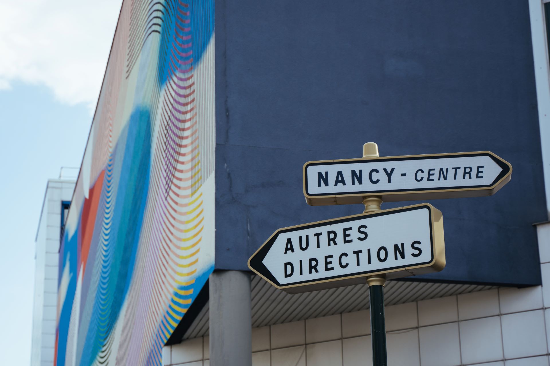 Deux panneaux routiers indiquent le chemin vers Nancy et d'autres directions. Derrière les panneaux se trouve un bâtiment peint en bleu foncé avec un mur coloré.