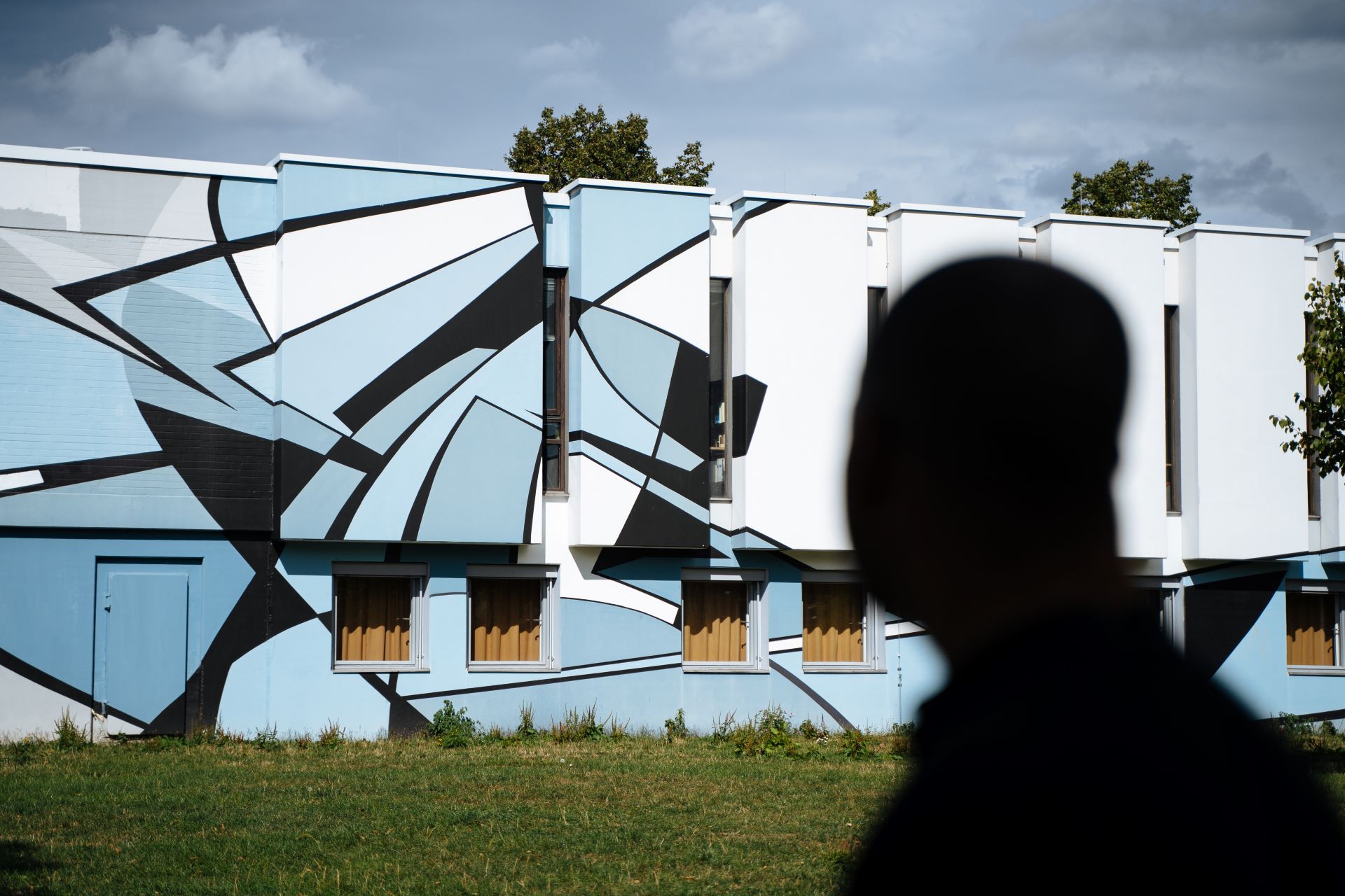 L'ombre d'un homme apparaît devant un bâtiment peint. Le bâtiment est peint avec du street art dans des tons de bleu.