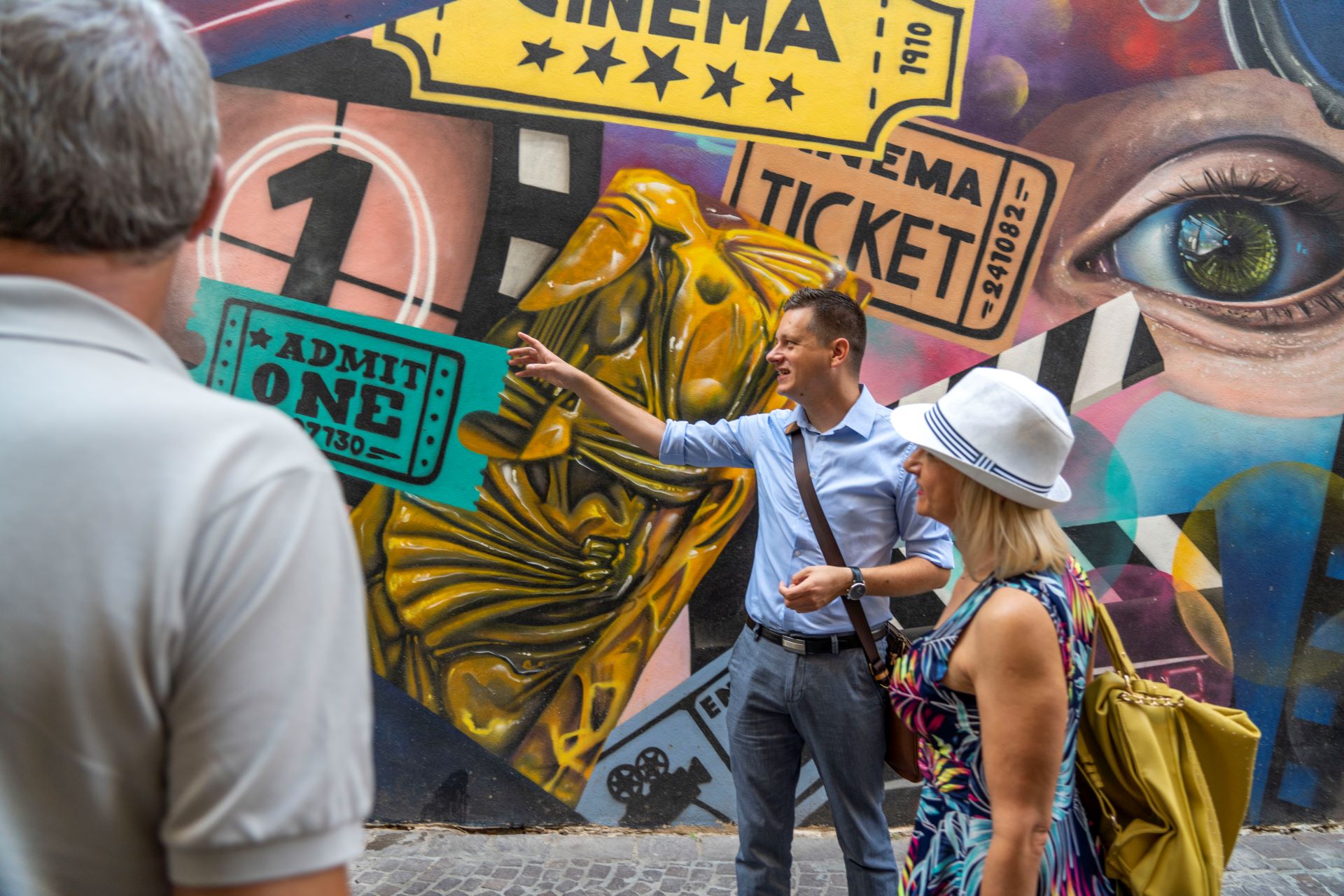 Un homme se tient devant un mur peint en couleur et montre quelque chose du doigt tout en expliquant à deux touristes.