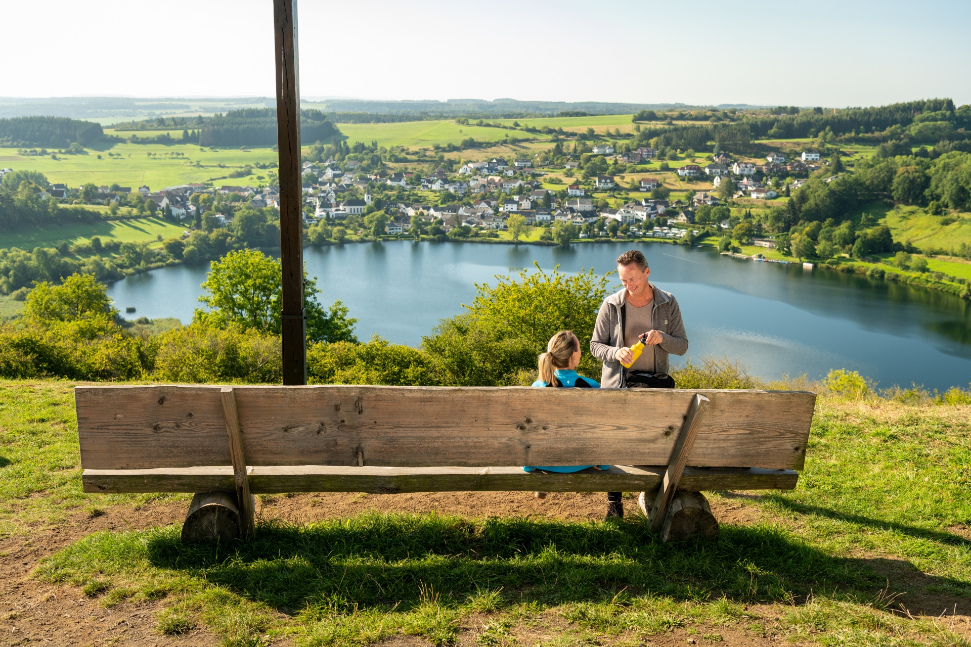Un couple en tenue de randonnée fait une pause au Maarkreuz avec une vue sur le Schalkenmehrener Maar