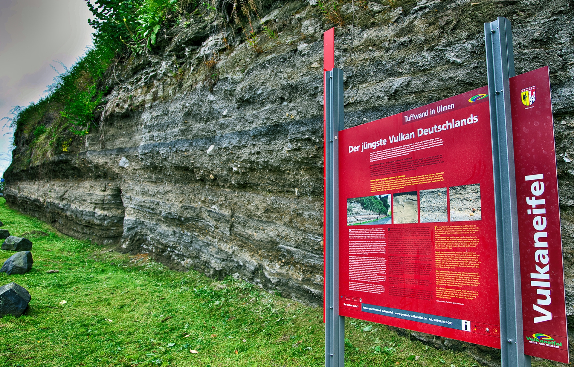 Un panneau d'information rouge destiné aux randonneurs se trouve devant un mur de tuf à Ulmen.