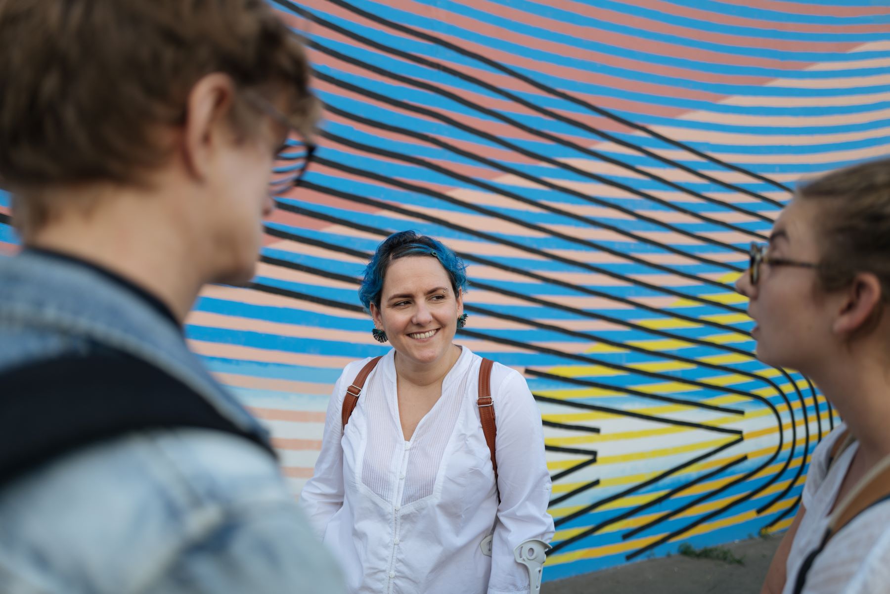 La guide touristique discute avec ses invités devant un grand mur peint à Nancy. La guide a des cheveux bleus qui s'accordent avec les tons bleus du mur derrière elle.