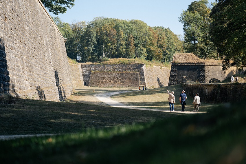 Les trois personnes marchent à l'intérieur de la forteresse en suivant les chemins tracés. Les chemins sont bordés de prairies vertes. Les trois personnes marchent le long d'un pont et visitent la fortification.