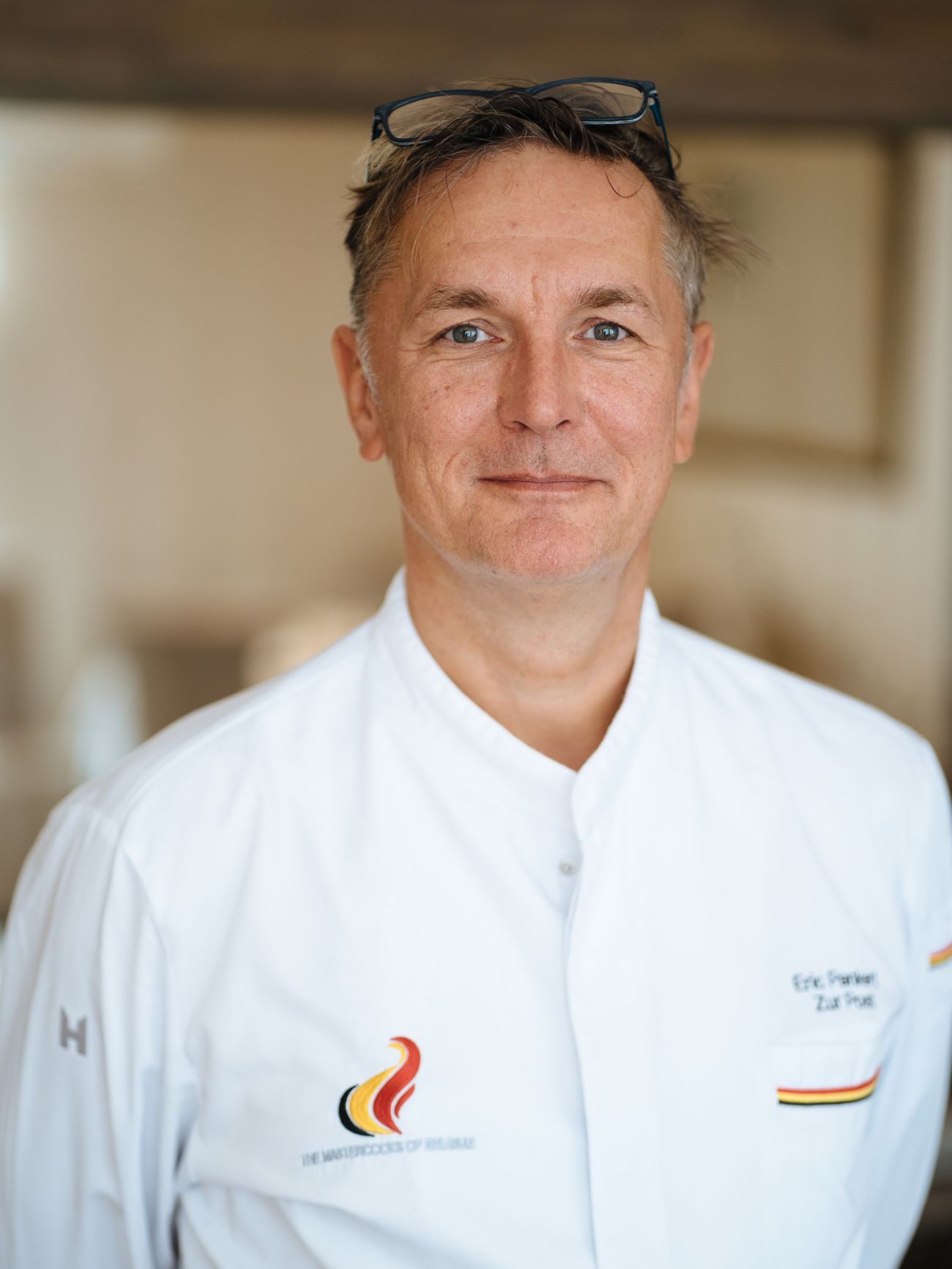 Dies ist ein Porträt von Erik Pankert, dem Chefkoch, der mit einem leichten Lächeln direkt in die Kamera schaut. Er hat graue Haare, trägt seine Brille auf dem Kopf und eine weiße Küchenuniform.