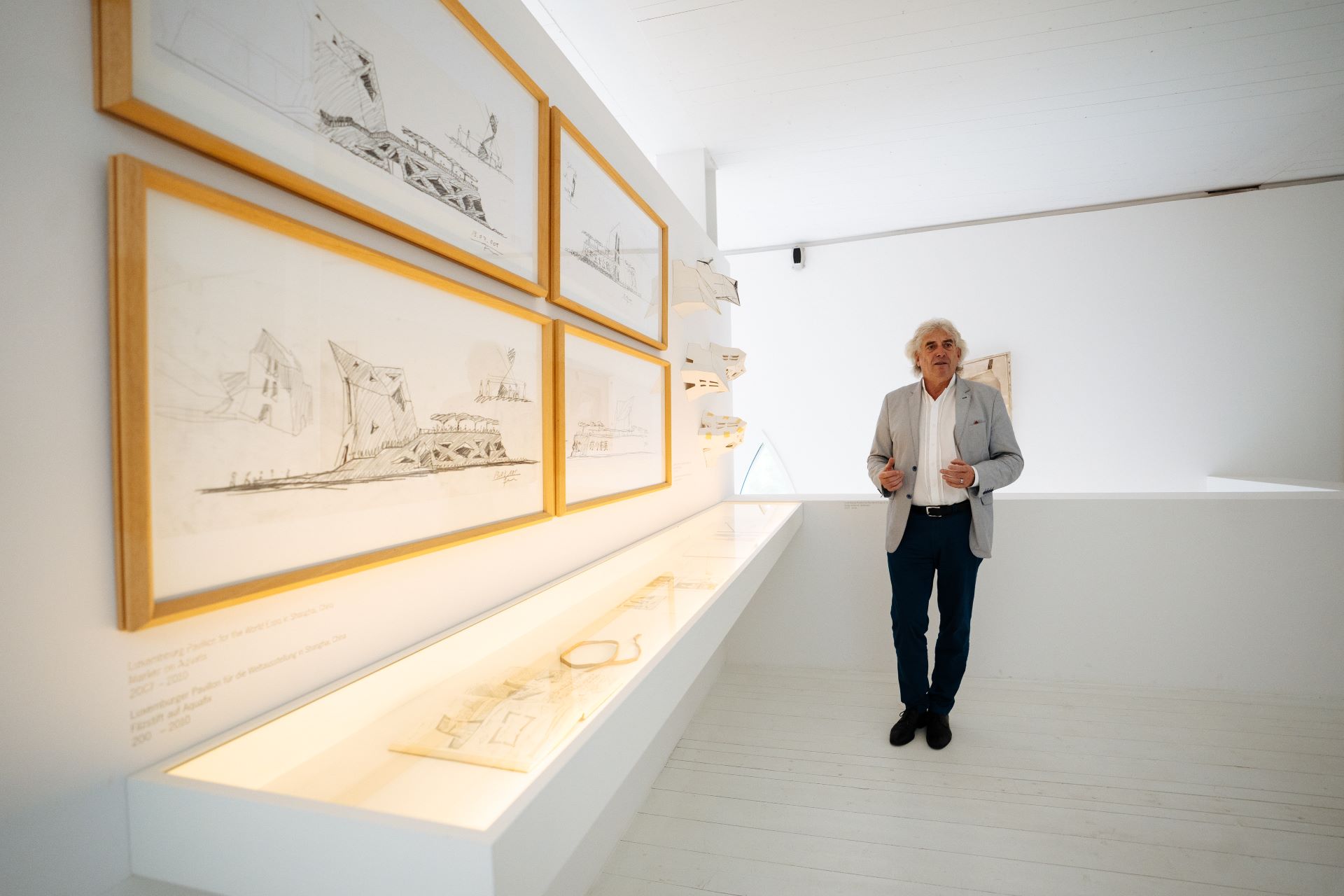 Valentiny geht vor einer architektonischen Kunstausstellung in einem komplett weißen Raum in einem Museum. Er trägt eine graue Anzugsjacke, ein weißes Hemd und eine dunkle Hose.