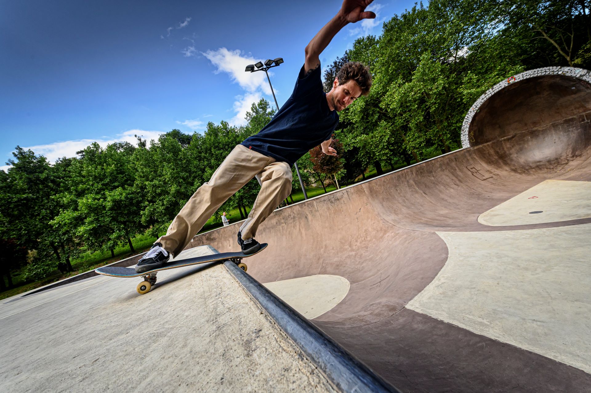 Ein Skateboarder macht einen Trick, indem er sich mit seinem Skateboard fast senkrecht auf den Boden stützt. Er befindet sich im Skatepark mit Bäumen und einem blauen Himmel im Hintergrund.