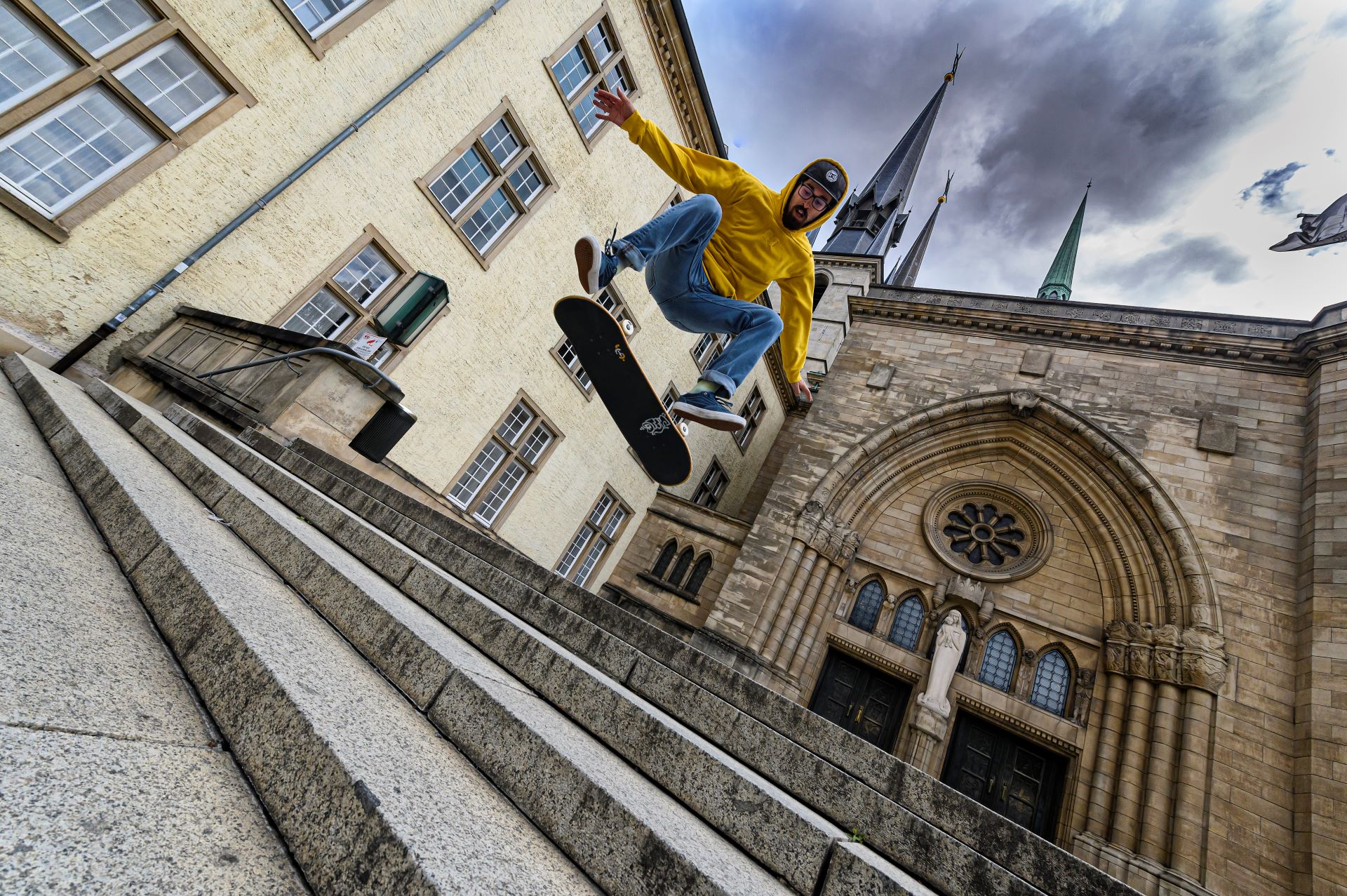 Ein Skateboarder in gelbem Kapuzenpulli und blauer Jeans macht einen Trick auf einer Treppe vor einer Kathedrale in Luxemburg.