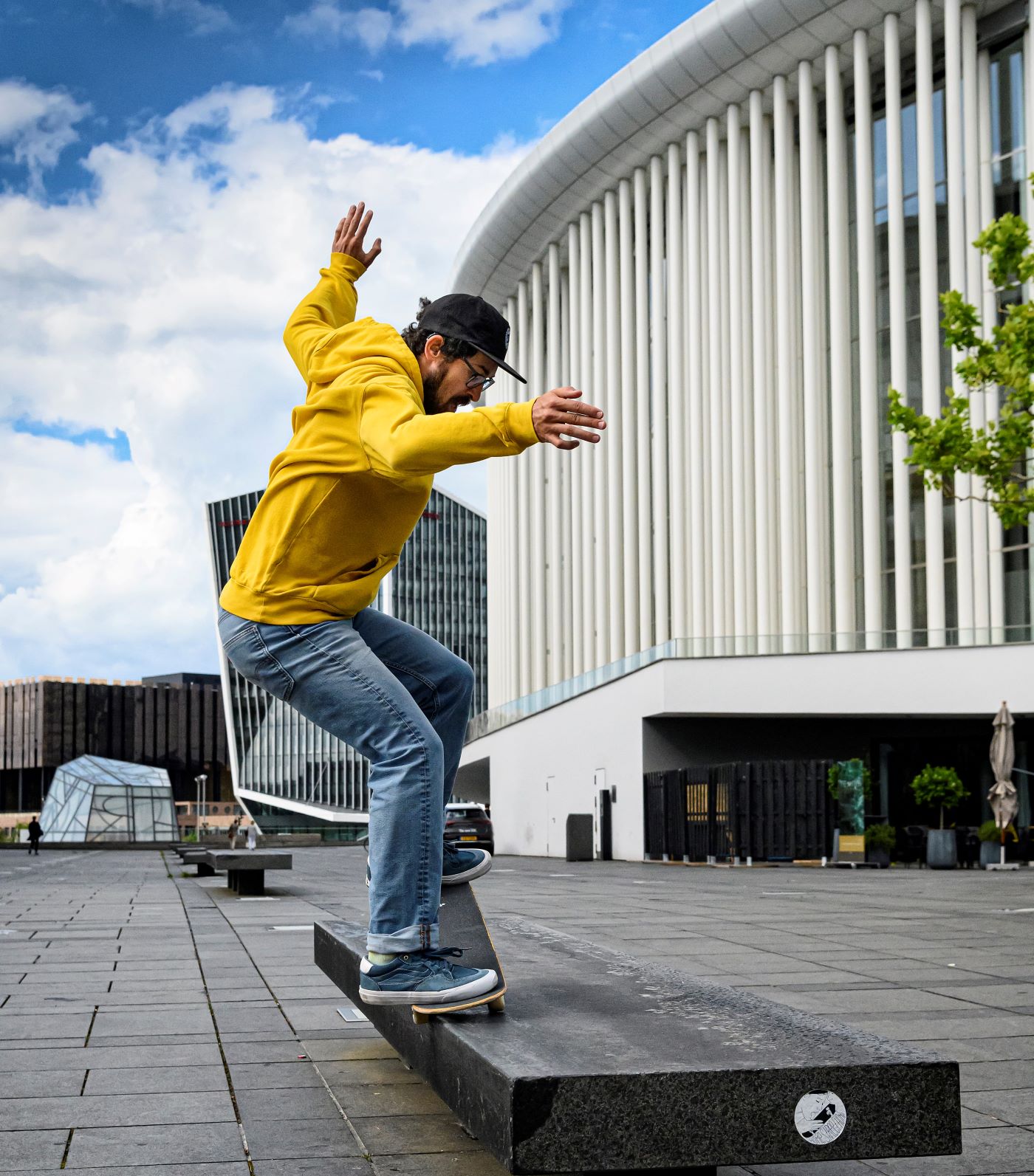 Ein Skateboarder mit gelbem Kapuzenpulli skatet am Rand einer Bank vor einem großen Gebäude entlang. Seine Hände sind in der Luft.