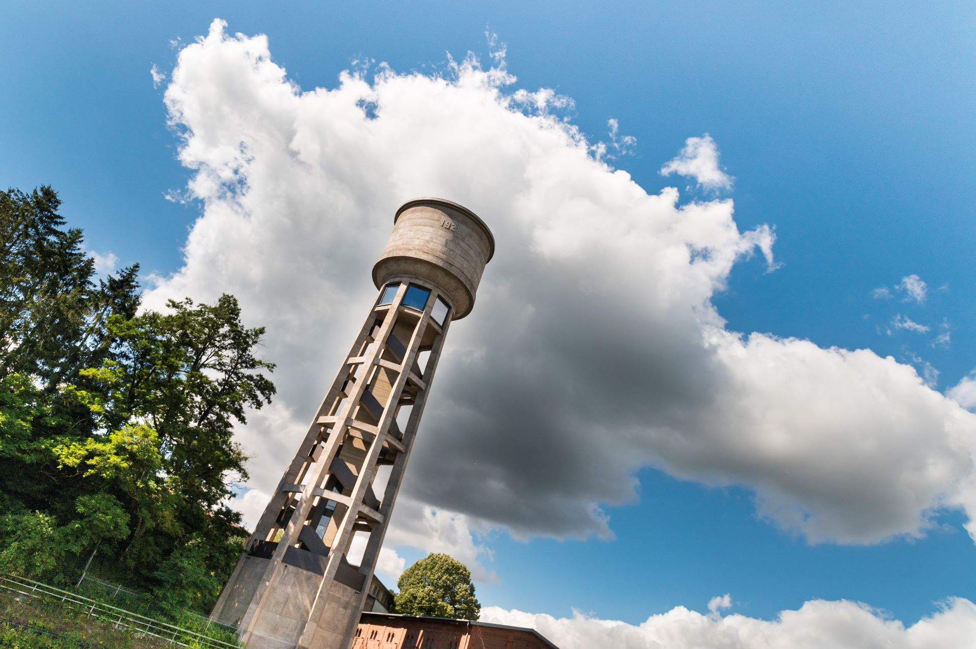Ein Wasserturm, der eine der Steichen-Fotokollektionen beherbergt, ist vor einem blauen Himmel mit weißen Wolken abgebildet. Im Hintergrund sind Bäume zu sehen.