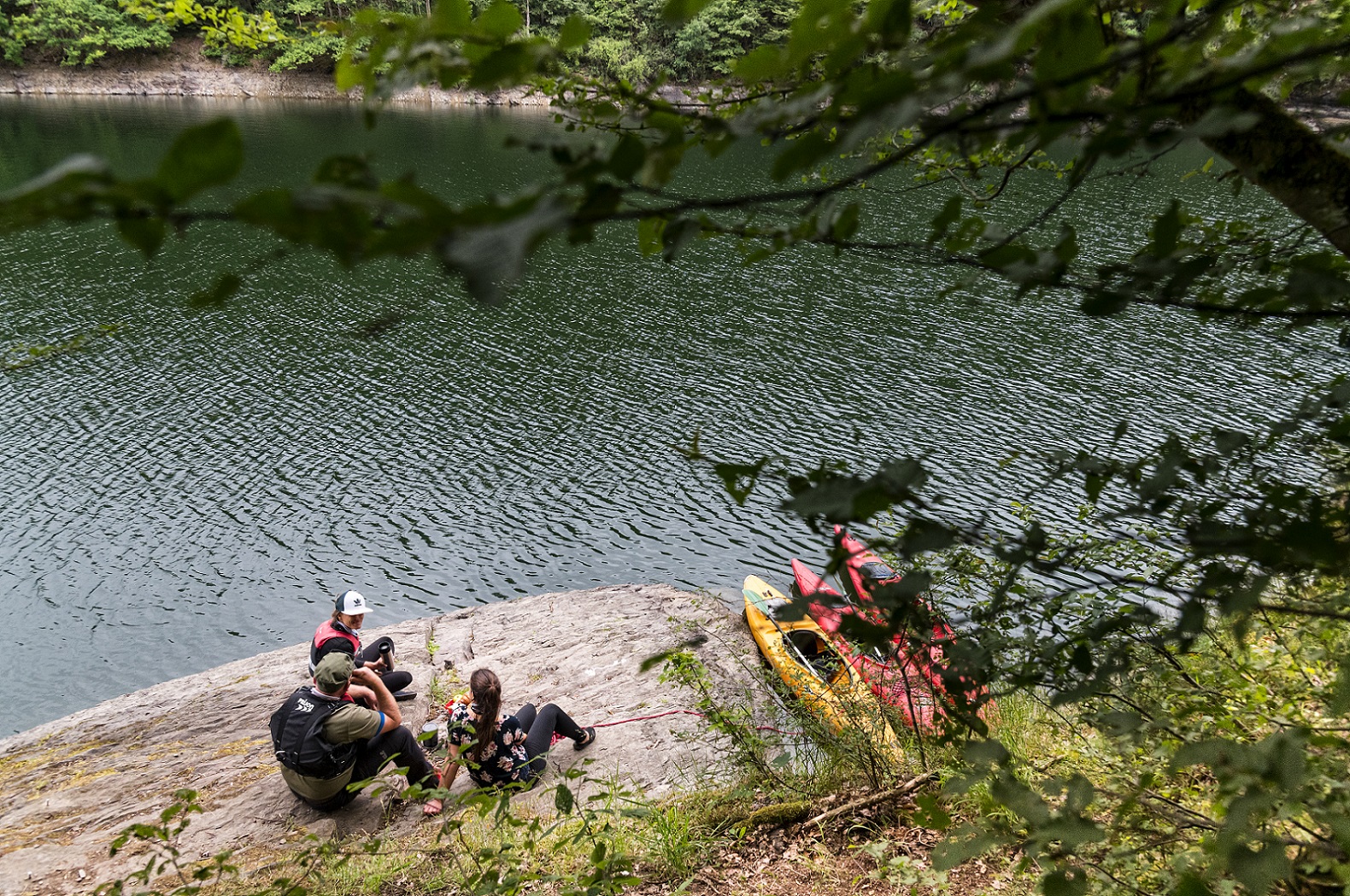 Eine Gruppe von drei Menschen machen eine Rast auf einem Felsen am Ufer des Stausees. Ihre Kayake liegen im Wasser.