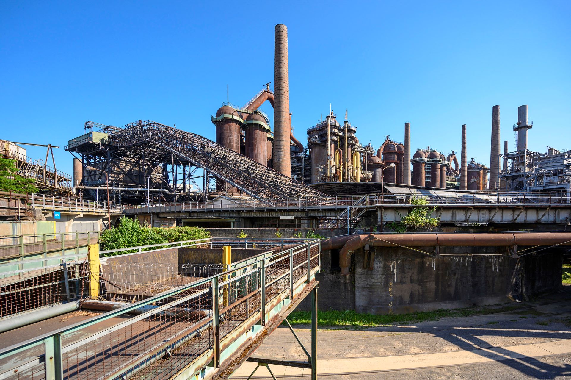 Im Hintergrund sind die rostfarbenen Strukturen, die die Völklinger Hütte ausmachen, abgebildet. Eine Industrieszene mit rostfarbenem Metall an verschiedenen historischen Maschinen ist vor blauem Himmel abgebildet.