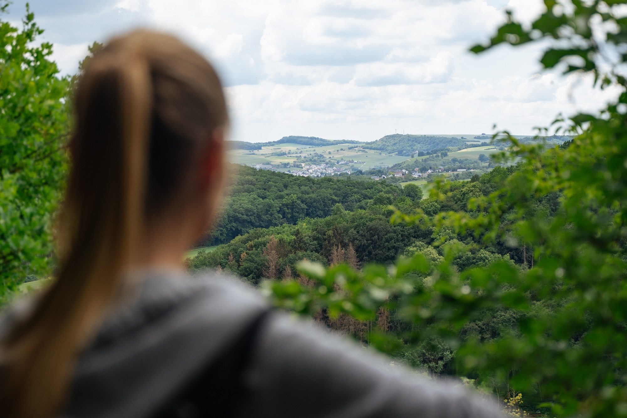 Landschaftsaufnahme des Blicks vom Plateau ins Tal. Im Vordergrund eine Teilnehmerin der Wanderung verschwommen in der Rückenansicht.