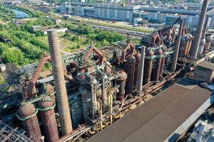 Ein Blick aus der Vogelperspektive auf das Industriegelände der Völklinger Hütte zeigt rostfarbene Rohre, große Gebäude und eine kleine Grünfläche im Hintergrund.