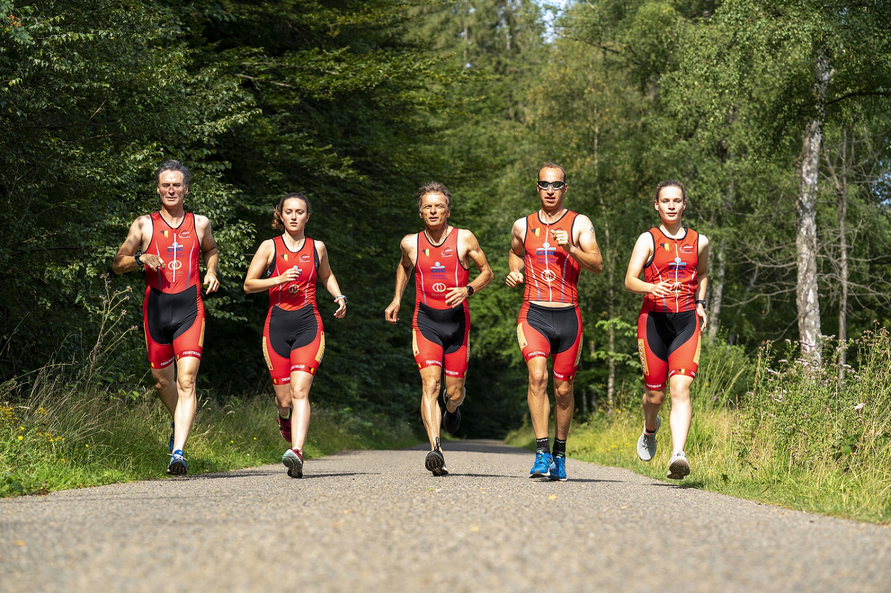Fünf Triathleten, darunter zwei junge Mädchen und drei ältere Herren, Laufen im Wald nebeneinander auf einem asphaltierten Weg.