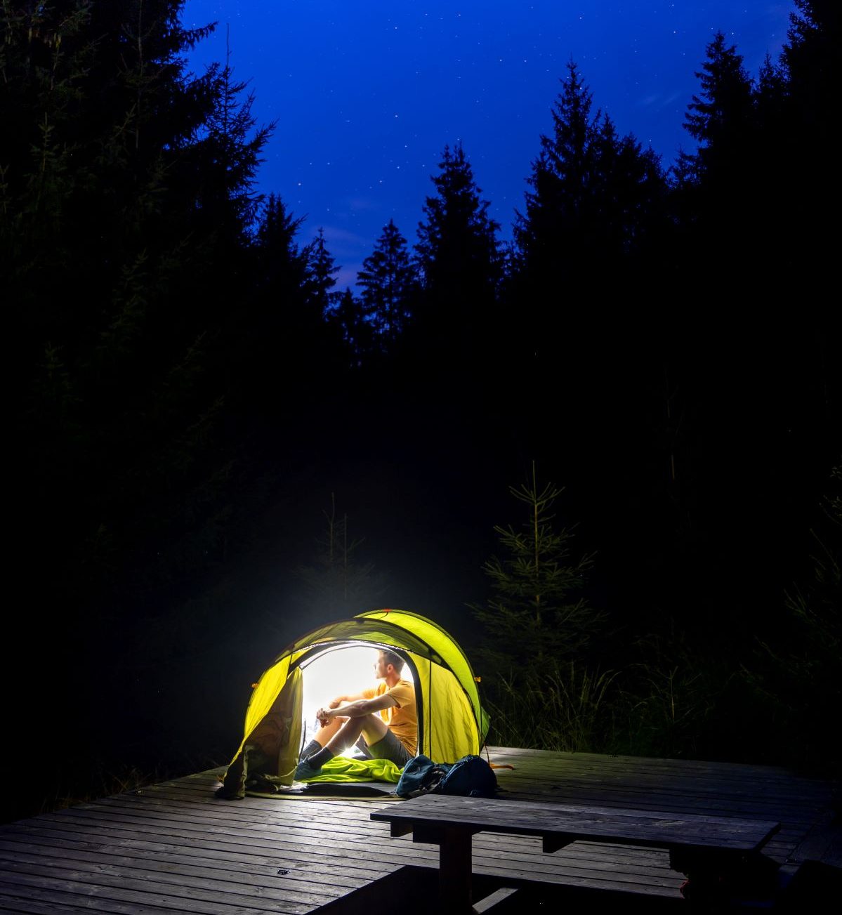Ein Mann mit einem gelben Hemd sitzt nachts in einem kleinen Zelt auf einer Holzplattform. Das Zelt ist beleuchtet und hell, aber der Himmel und die umliegenden Wälder sind dunkel.