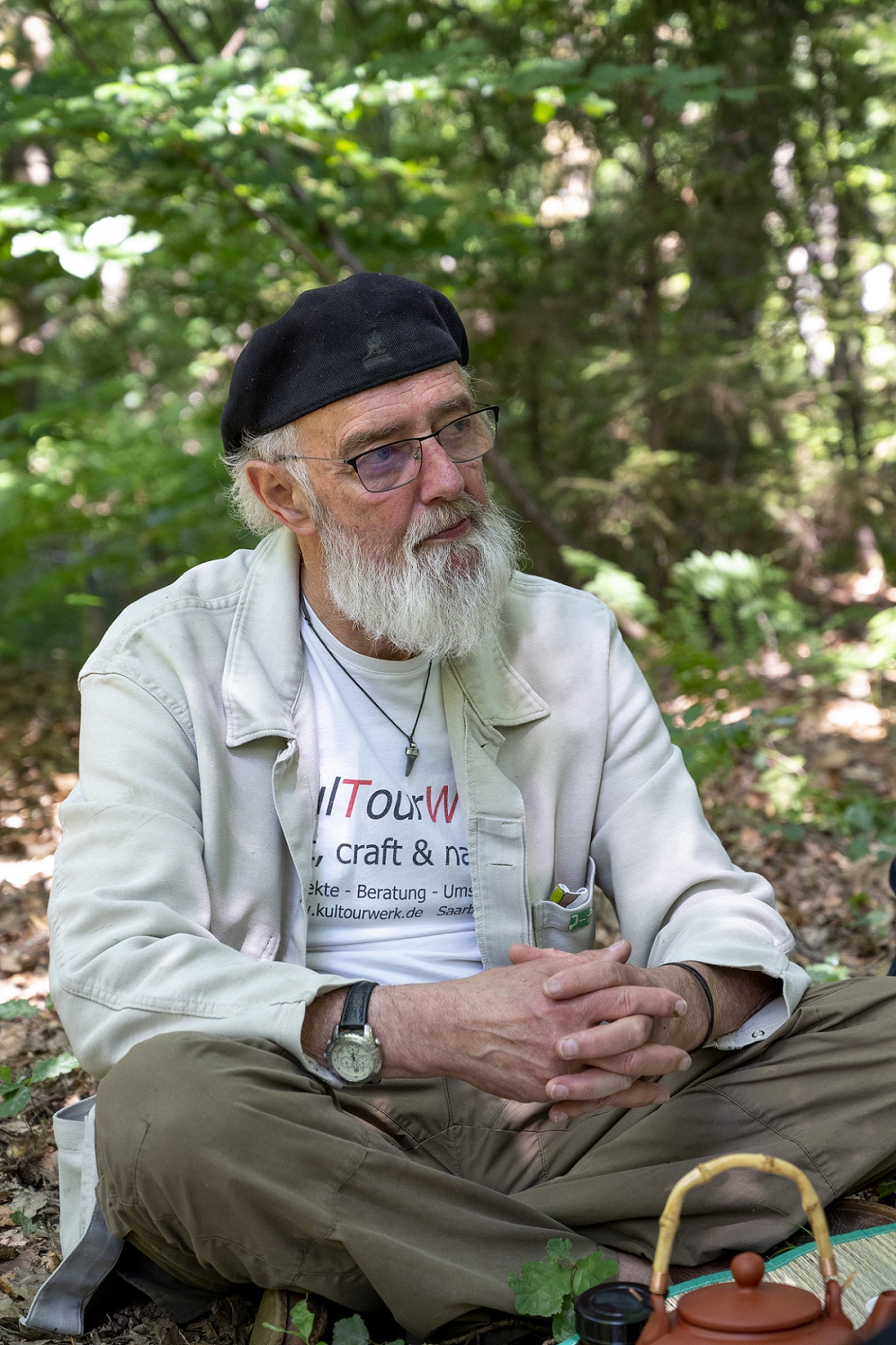 Portraitaufnahme von Lothar Wilhelm sitzend auf dem Waldboden mit ineinander verschränkten Händen. Er hat den Blick von der kamera abgewandt.