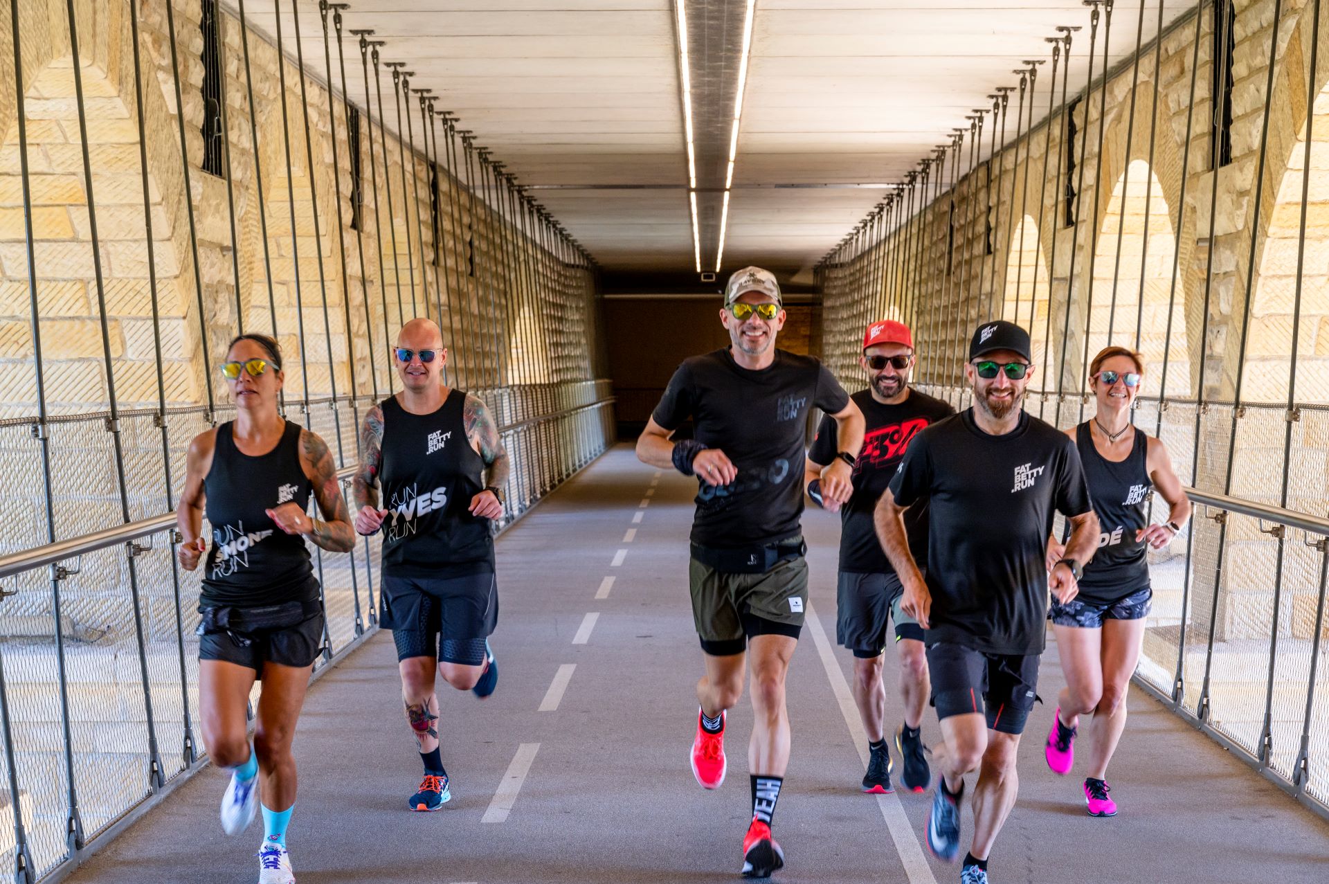 Sechs Mitglieder des Laufteams von FatBetty.run sind auf dieser Aktionsaufnahme zu sehen. Die Läufer überqueren eine überdachte Brücke aus Stein, tragen dunkle Laufkleidung, Sonnenbrillen und ein breites Lächeln.