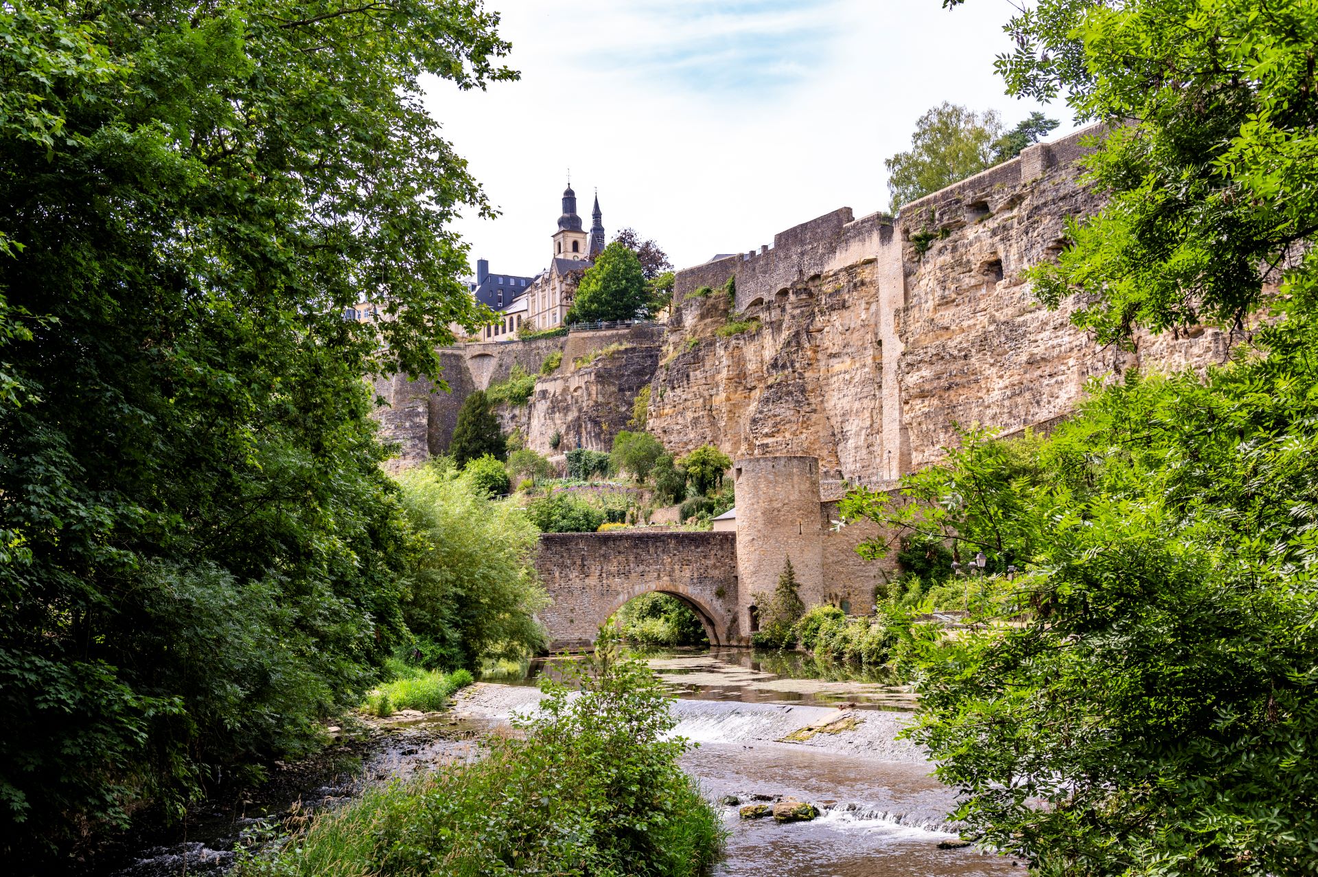Zwischen Bäumen bietet sich ein Blick auf die Luxemburger Festungsmauern und eine alte Steinbrücke. Der Stein ist beigefarben und die Bäume sind hellgrün, aber der Himmel ist kaum zu sehen.