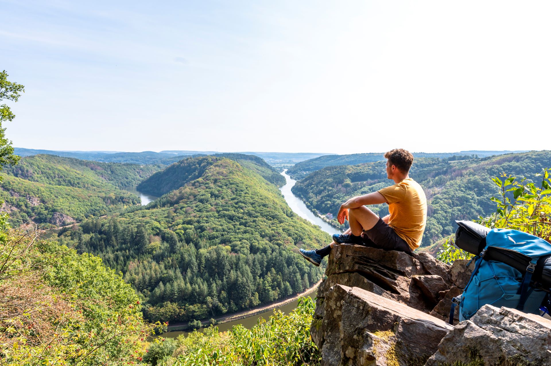 Ein Mann mit gelbem Hemd und Wanderausrüstung sitzt auf einem Felsvorsprung mit Blick auf die Saarschleife. Der Himmel ist blau und die umliegenden Wälder sind leuchtend grün.