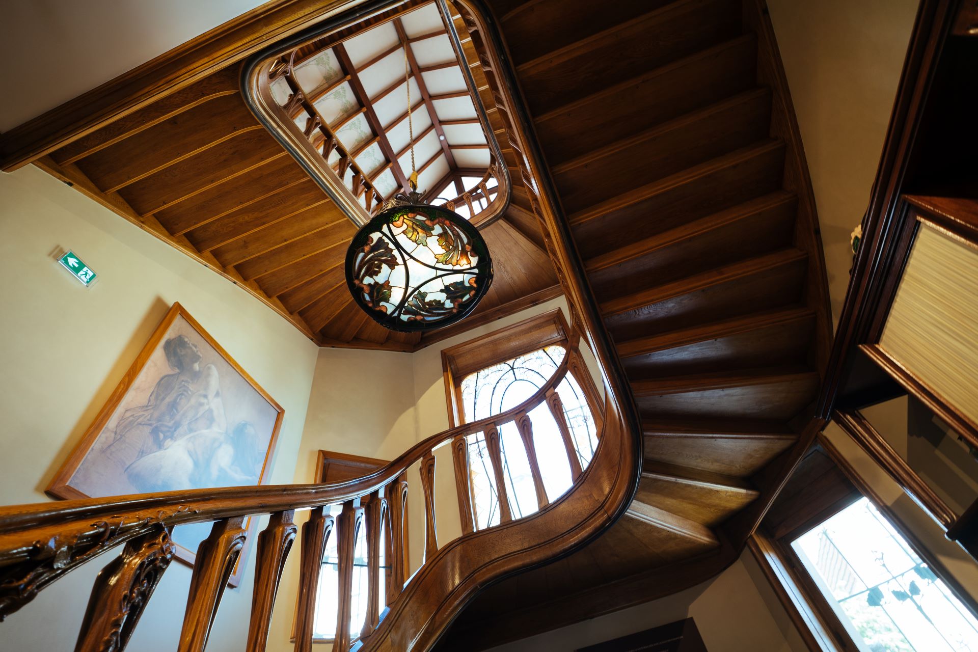 Von unten ist eine Holztreppe zu sehen, die den Kurven der Natur nachempfunden ist. In der Mitte des Bildes hängt eine Buntglaslampe.
