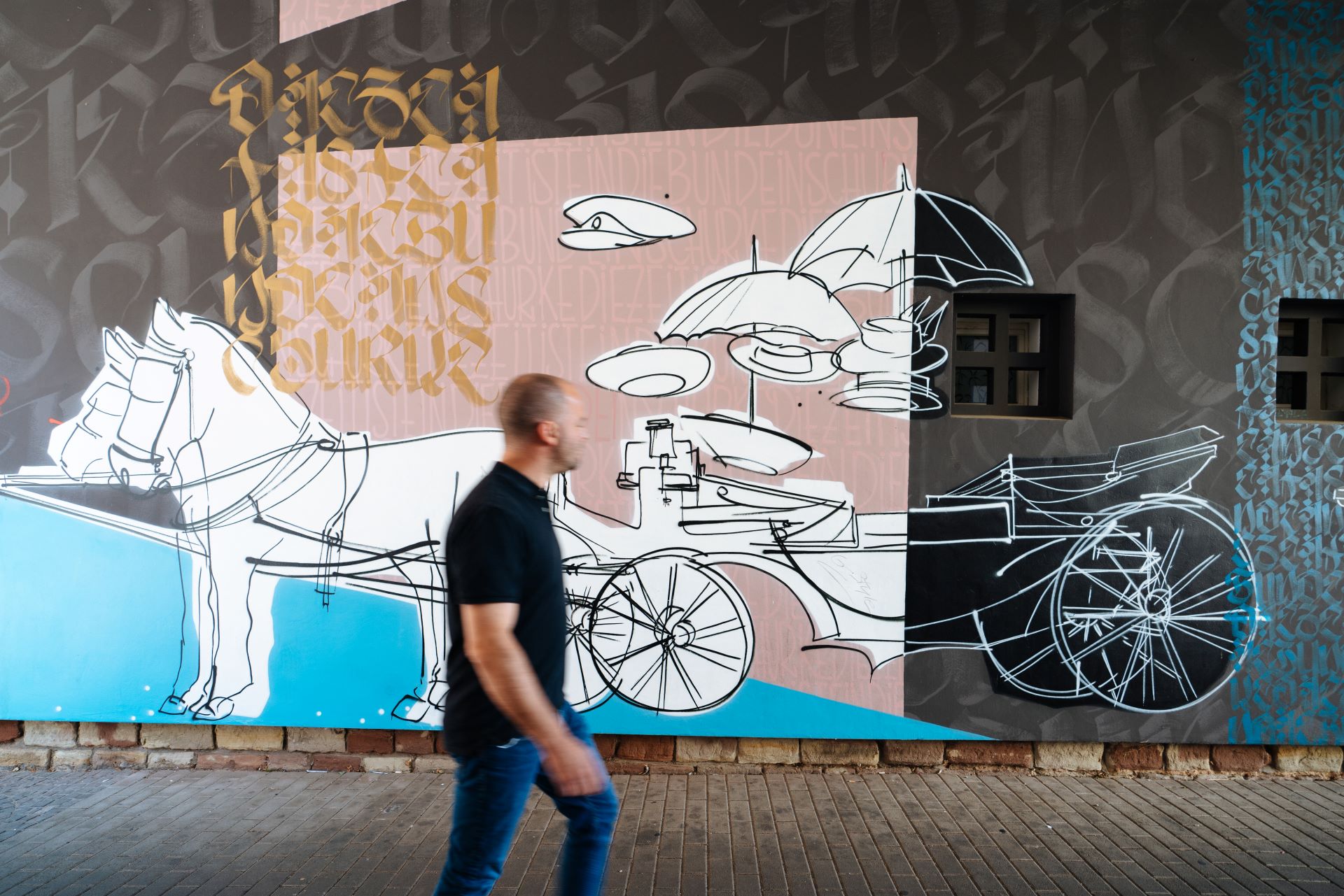 Reso geht vor einer mit Graffiti bemalten Wand. Das Kunstwerk stellt zwei Pferde dar, die eine Kutsche ziehen.
