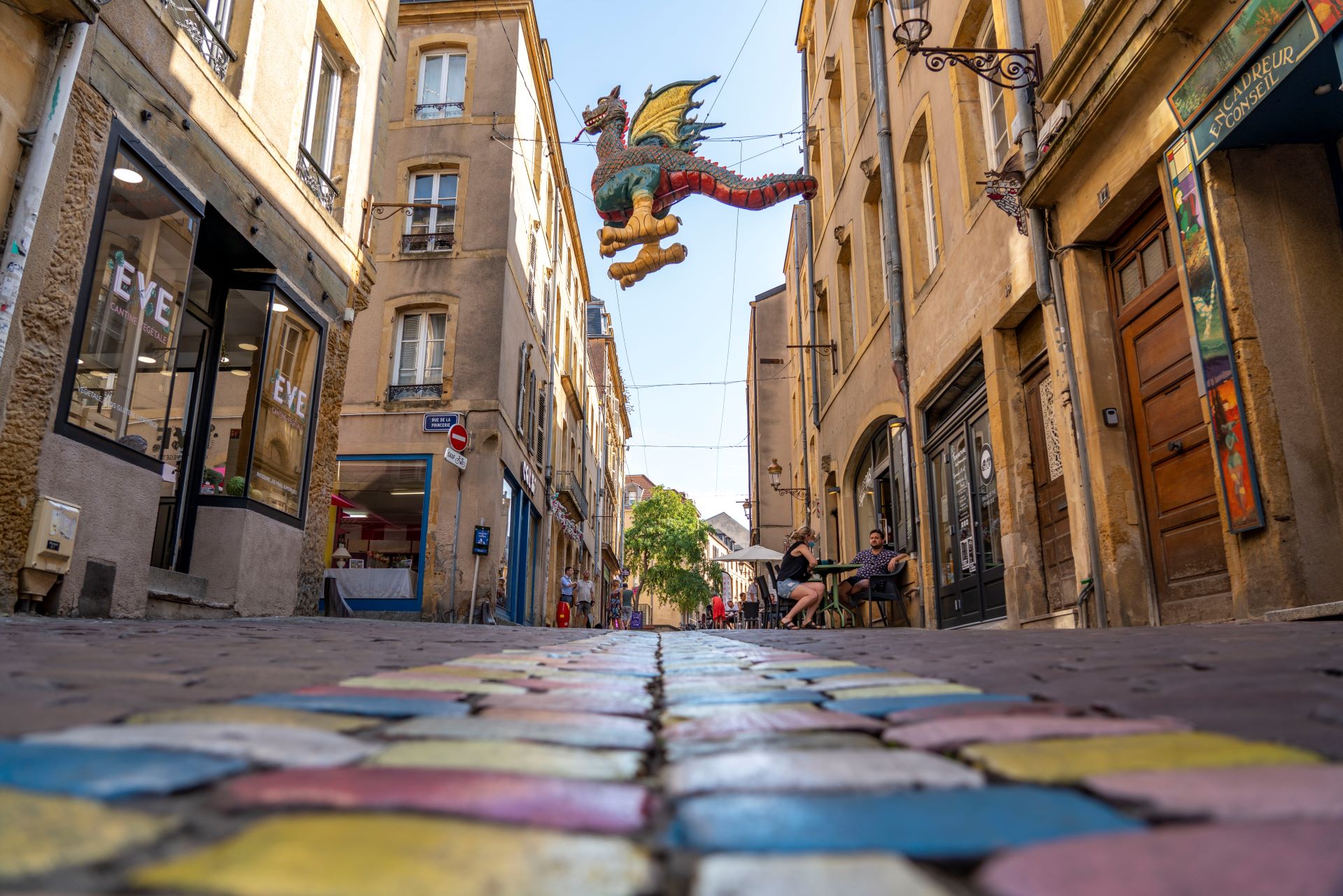 Der berühmte Drache von Metz schwebt über einer bunten Straße. Gelbe Gebäude und bemalte Straßen sorgen für eine verspielte Atmosphäre.