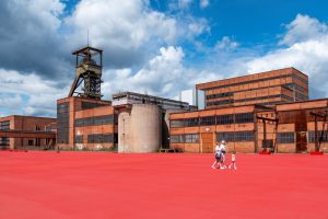 Die Familie geht über eine rot gestrichene Freifläche vor dem Bergwerk Wendel.