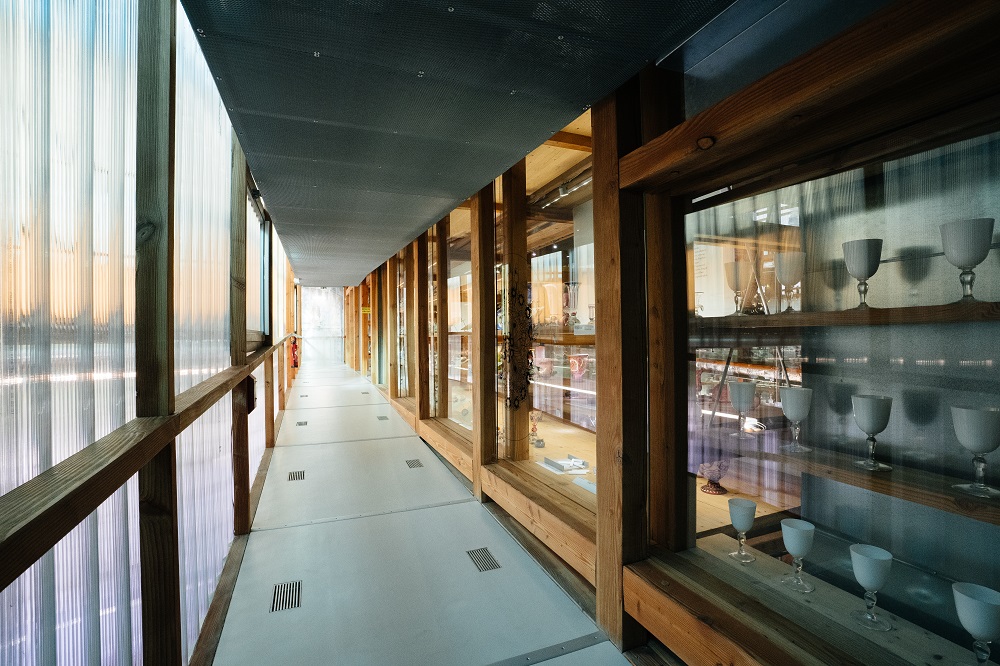 Aufnahme in einen Gang hinein. Links sind Fenster nach draußen zu sehen. Rechts entlang des Gangs befinden sich ausgestellte Glaskunstwerke hinter Fensterscheiben.