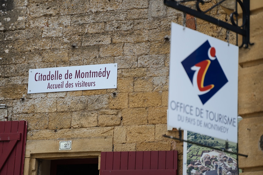 Im Vordergrund ist ein Schild zu sehen, dass die Touristeninformation von Montmedy ankündigt. Im Hintergrund ist scharf das Schild mit der Aufschrift "Citadelle de Montmédy - Accueil des visiteurs" zu lesen.